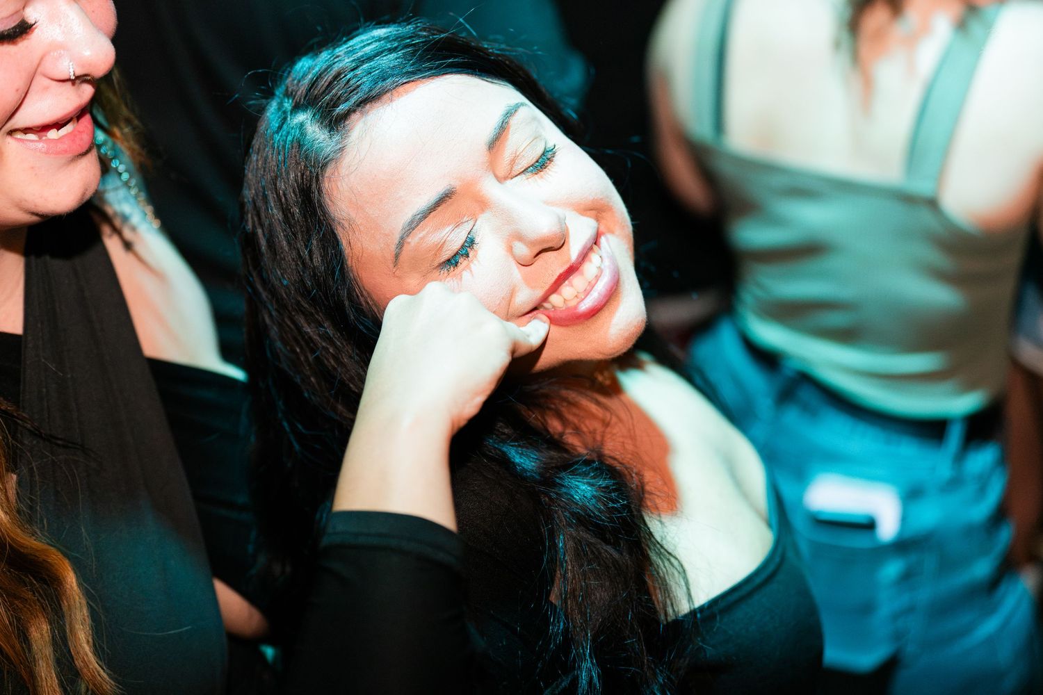 Woman smiling with eyes closed and hand on cheek under teal lighting at a nightclub.