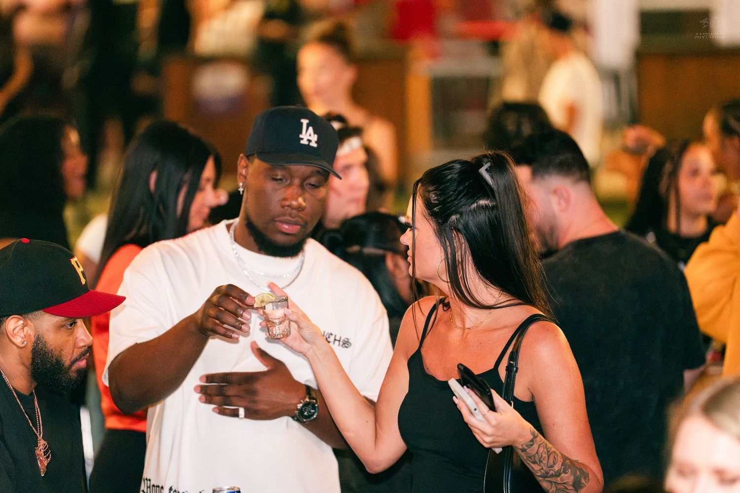 Man in LA cap and woman in black dress chatting at a lively social event.