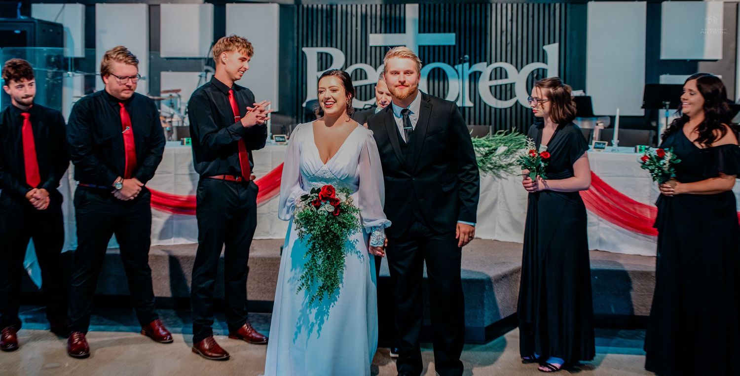 Wedding party poses together at indoor reception venue with red and white decorations and bouquet.