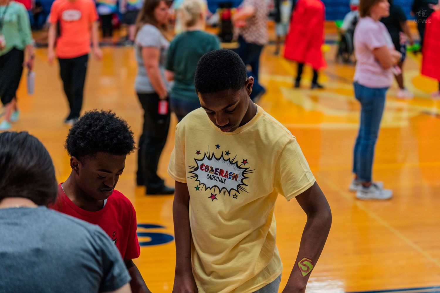 Students participate in an indoor gym activity, with a boy in a yellow shirt standing in the foreground.