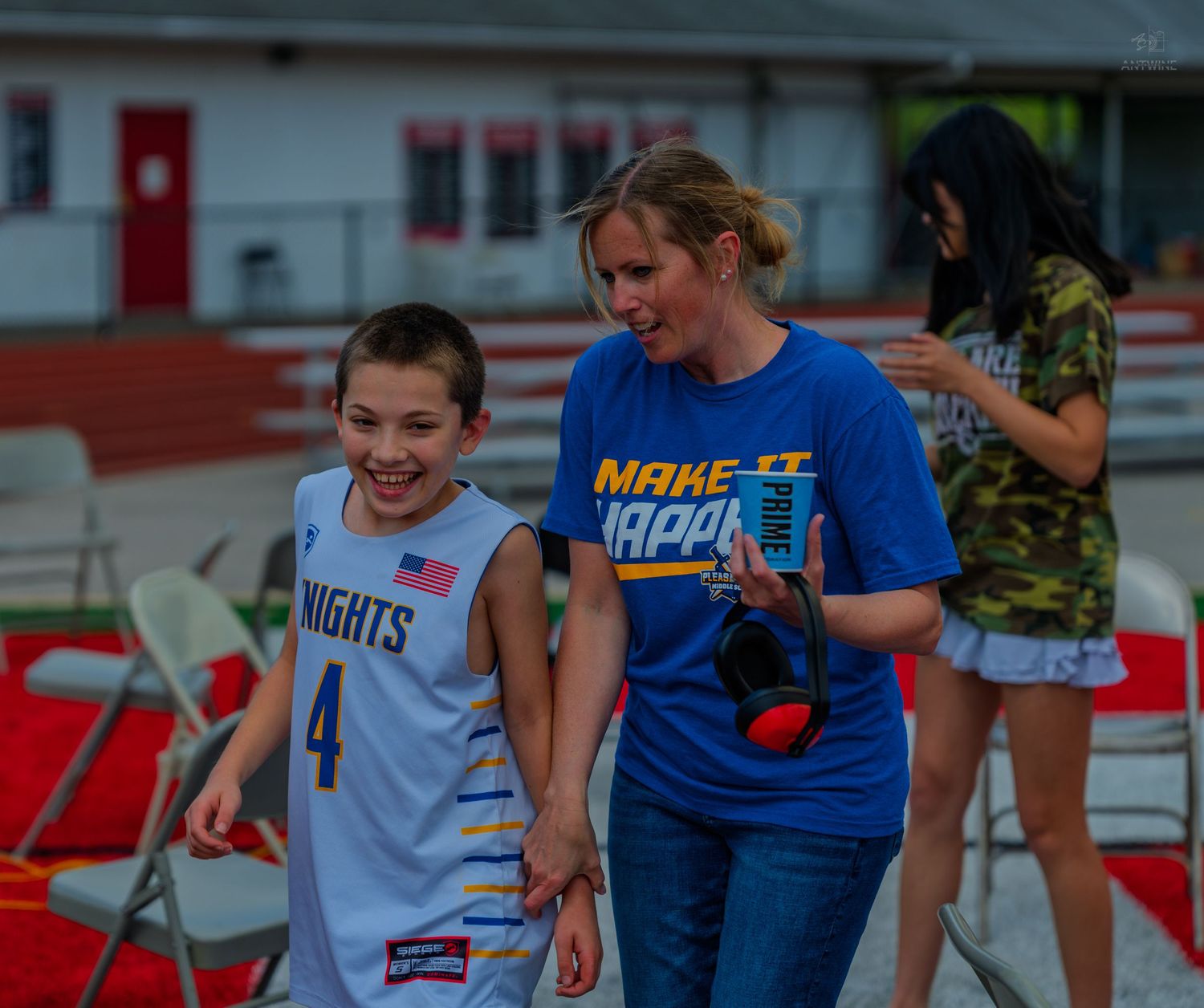 A woman in a blue Make It Happen shirt walks with a young boy in a Knights basketball jersey at an outdoor event.