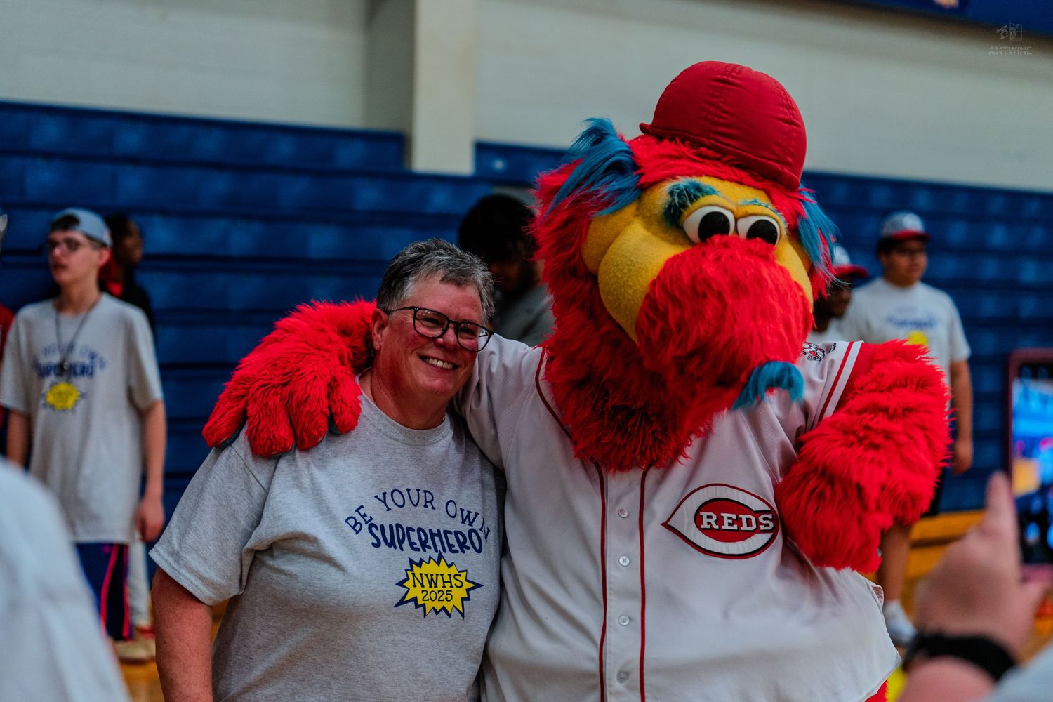 A woman poses with the Cincinnati Reds mascot Mr. Redlegs at an indoor sports event, both smiling for the camera.