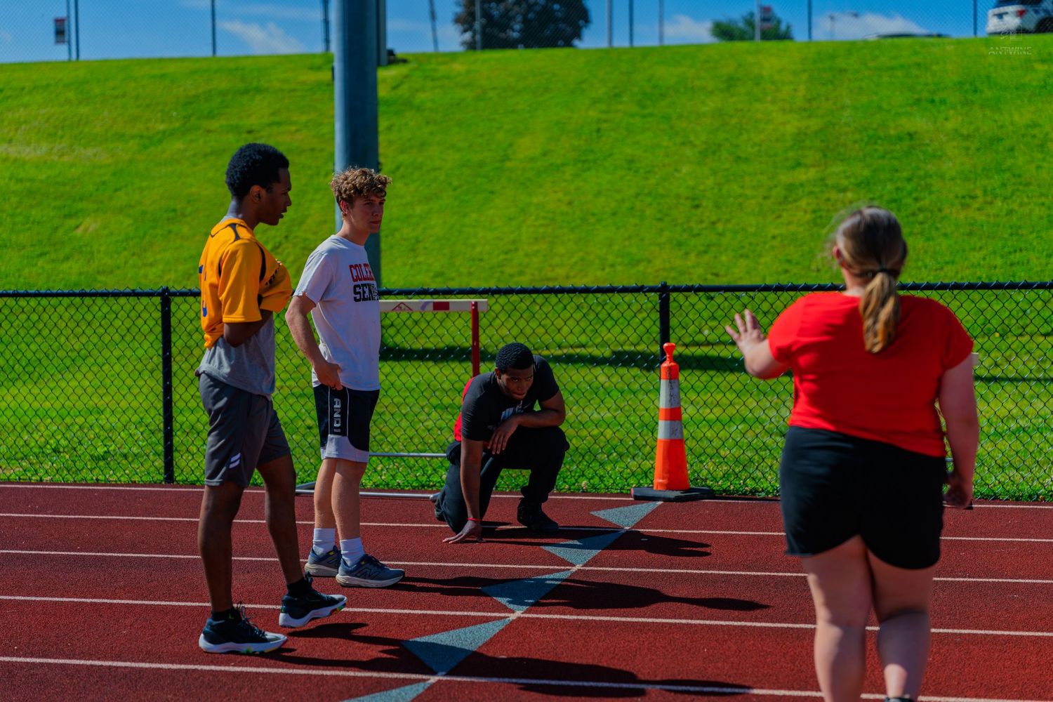 Coach instructs youth athletes on a red running track during outdoor sports practice on a sunny day.