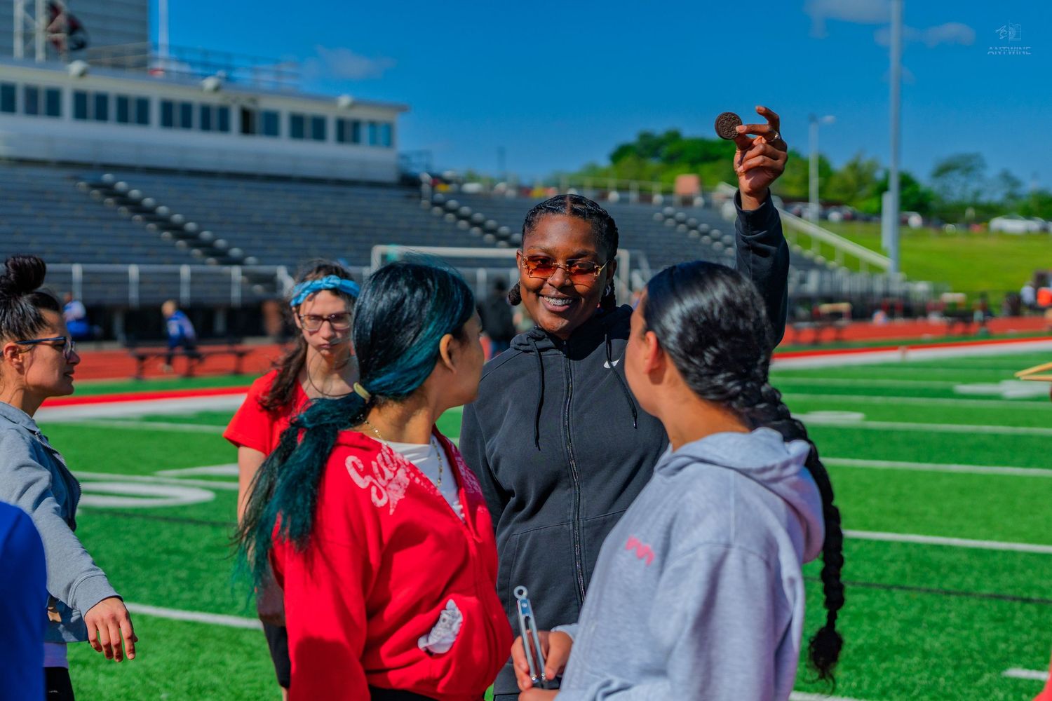 A group of students interact with a coach on a sunny athletic track field during an outdoor sports event.