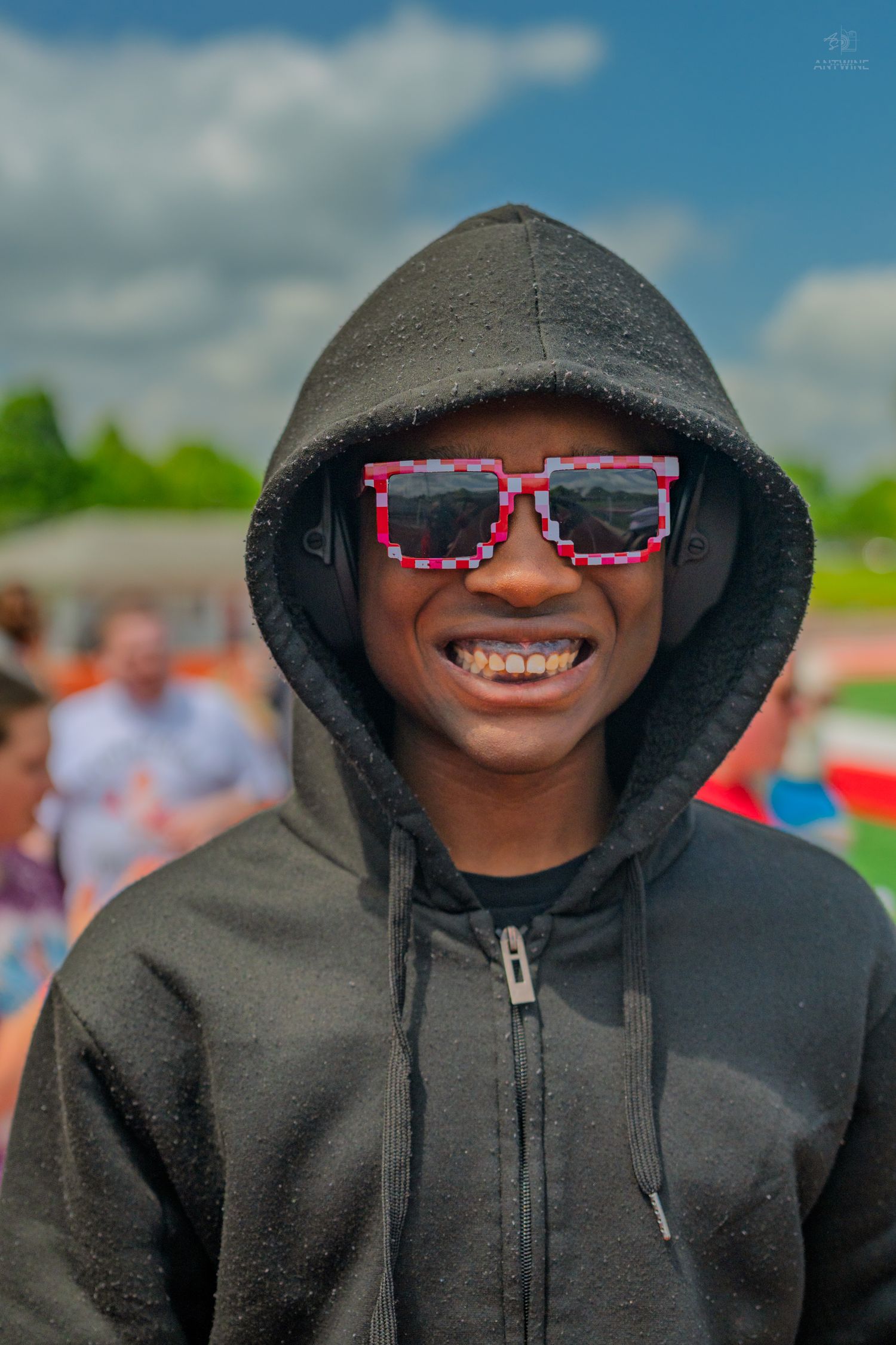 Smiling young man wearing a black hoodie and pixelated sunglasses outdoors on a sunny day.
