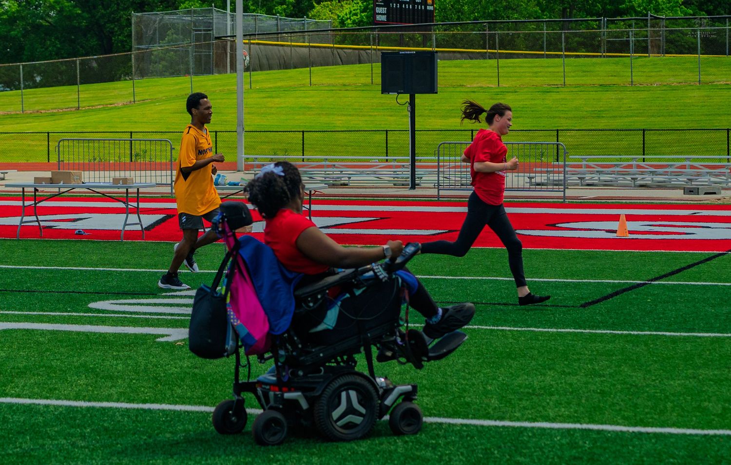 Person in motorized wheelchair watches runners on a red track at an outdoor athletic field during a track event.