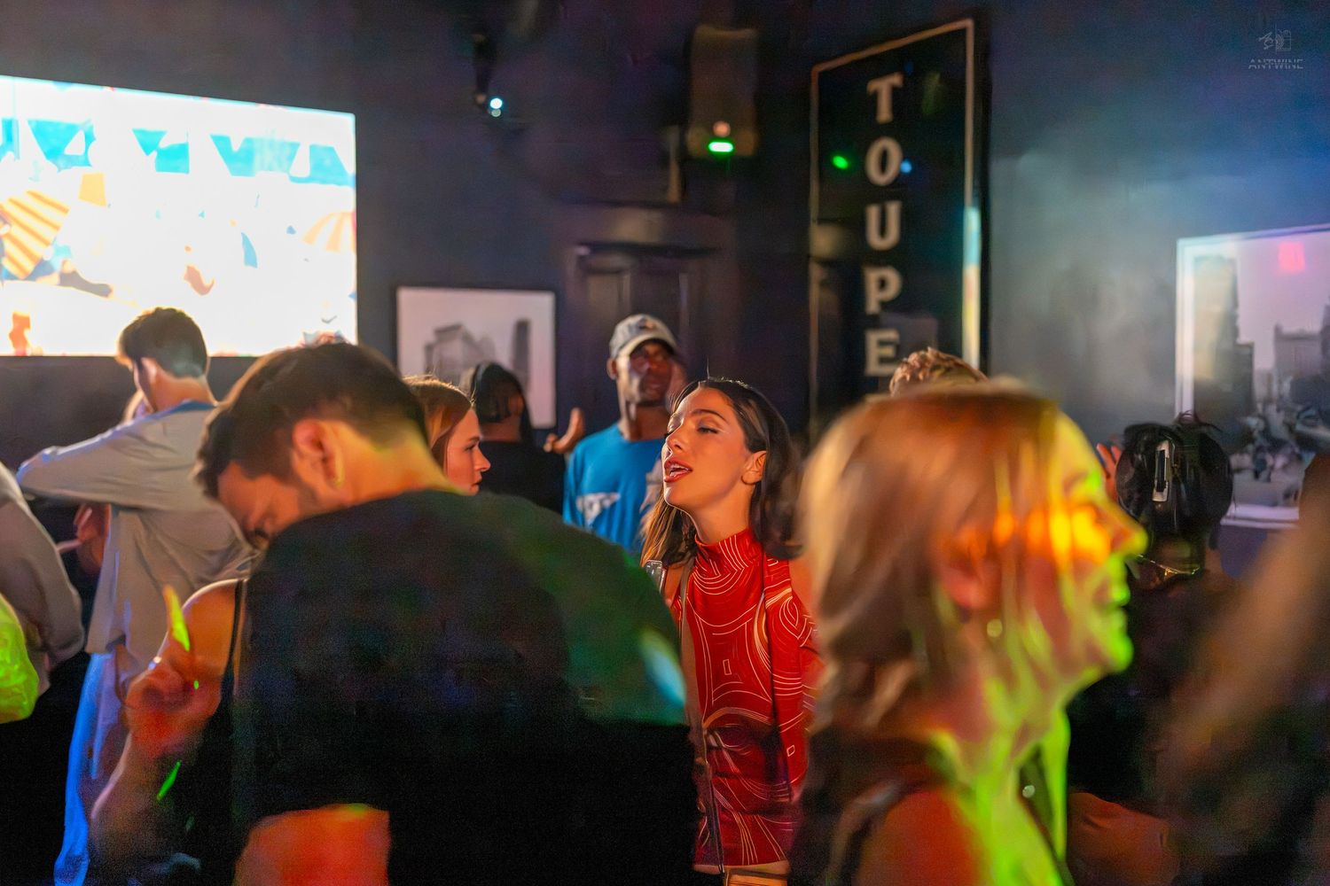 People dancing and socializing at a lively nightclub event with colorful lights and a projection screen.