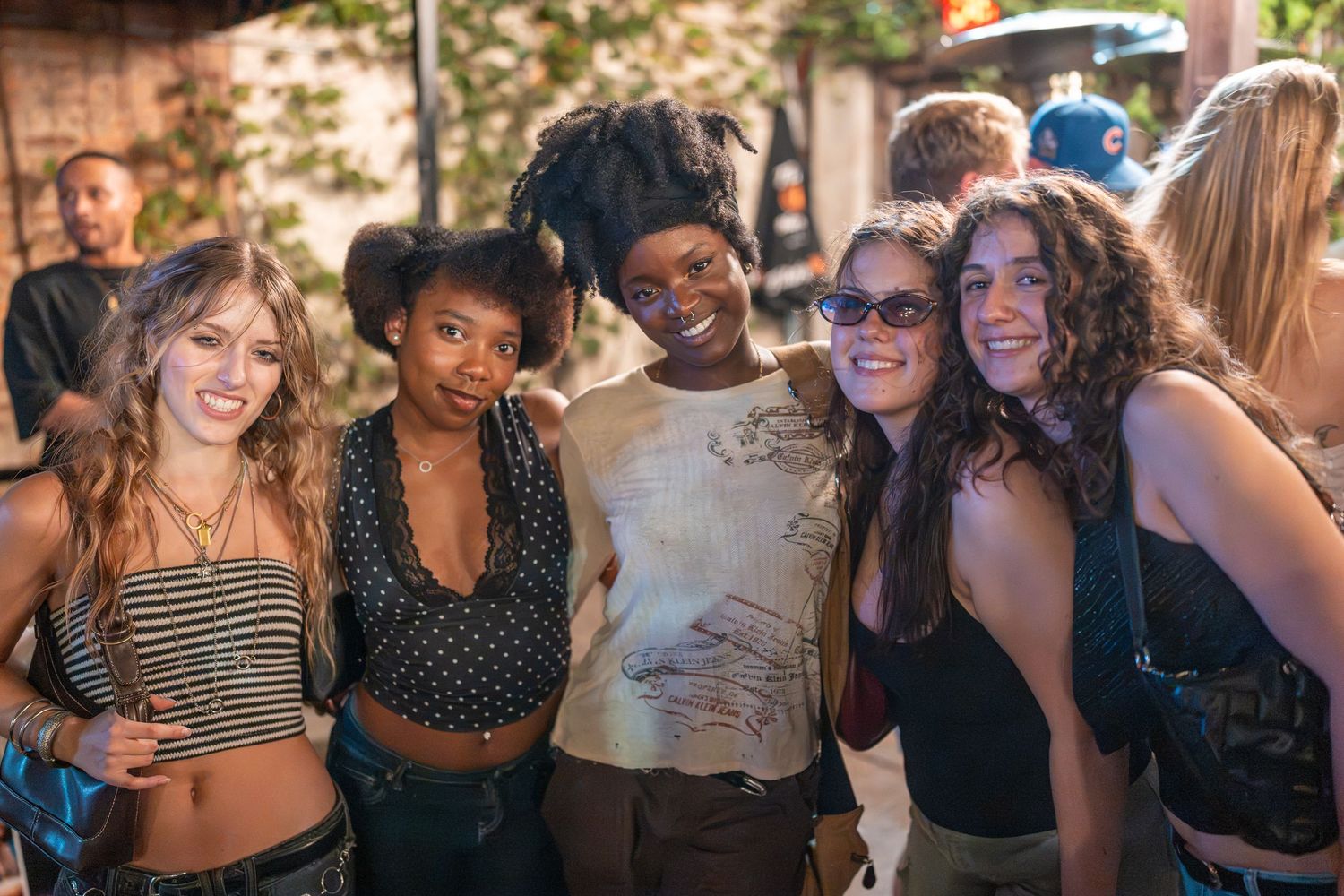 Five young women smiling together at an outdoor social gathering, posing for a group photo.