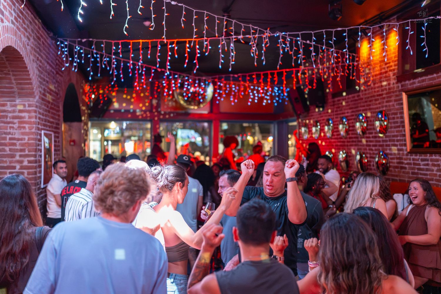 Crowded bar with string lights, brick walls, and people dancing and socializing at a lively indoor party.