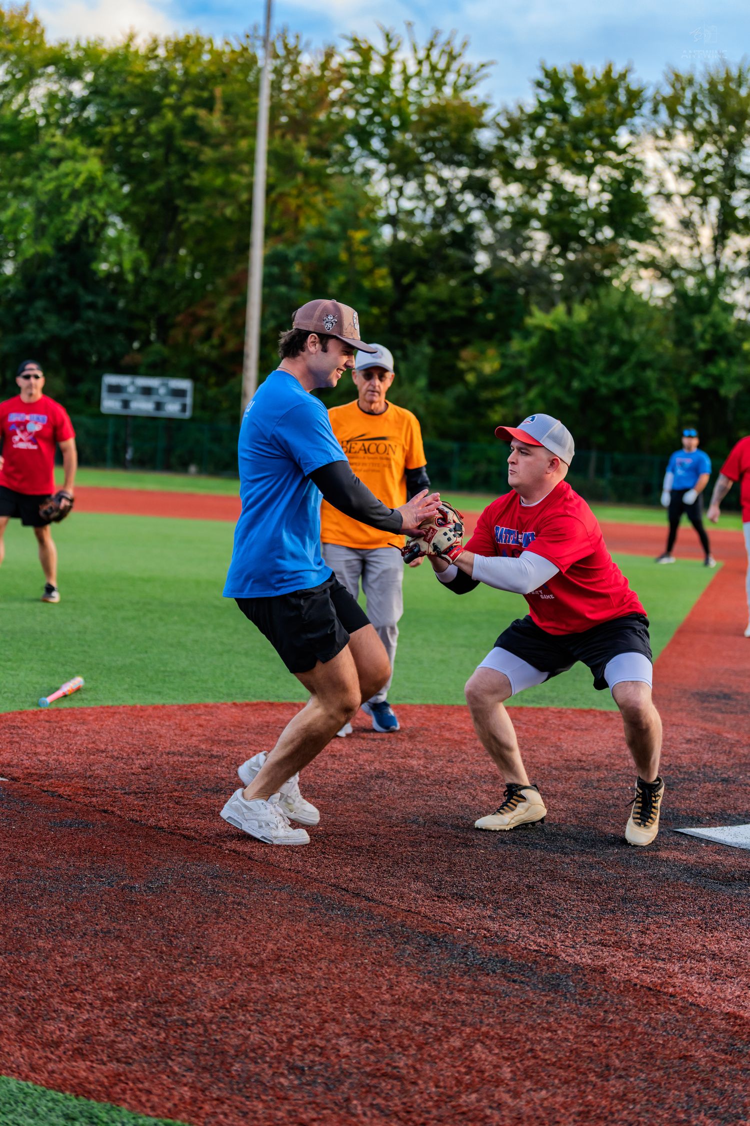 A baseball player in a red jersey tags out a runner in blue during a recreational softball game on a red turf field.