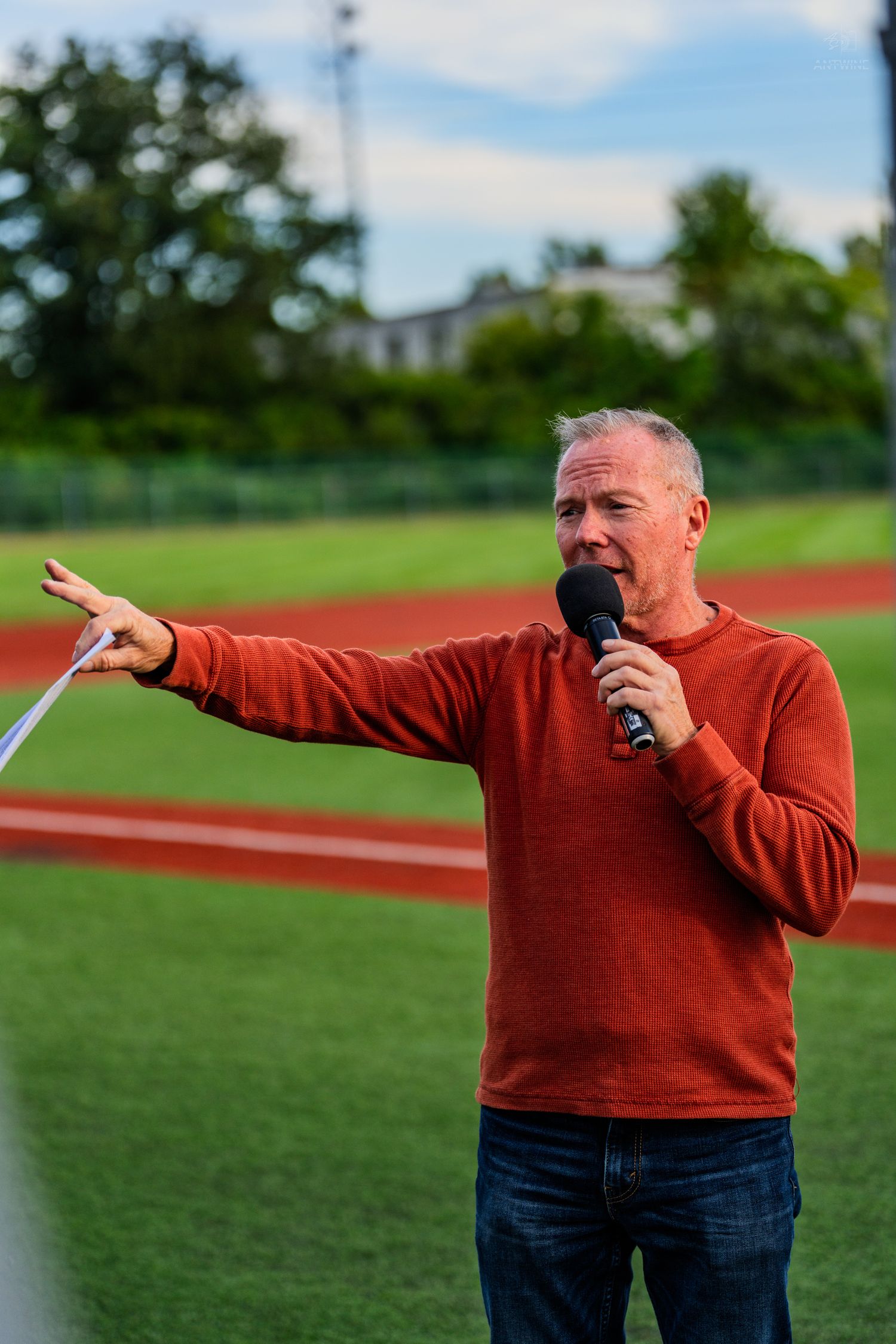 Man in red sweater speaking into microphone at outdoor baseball field event.