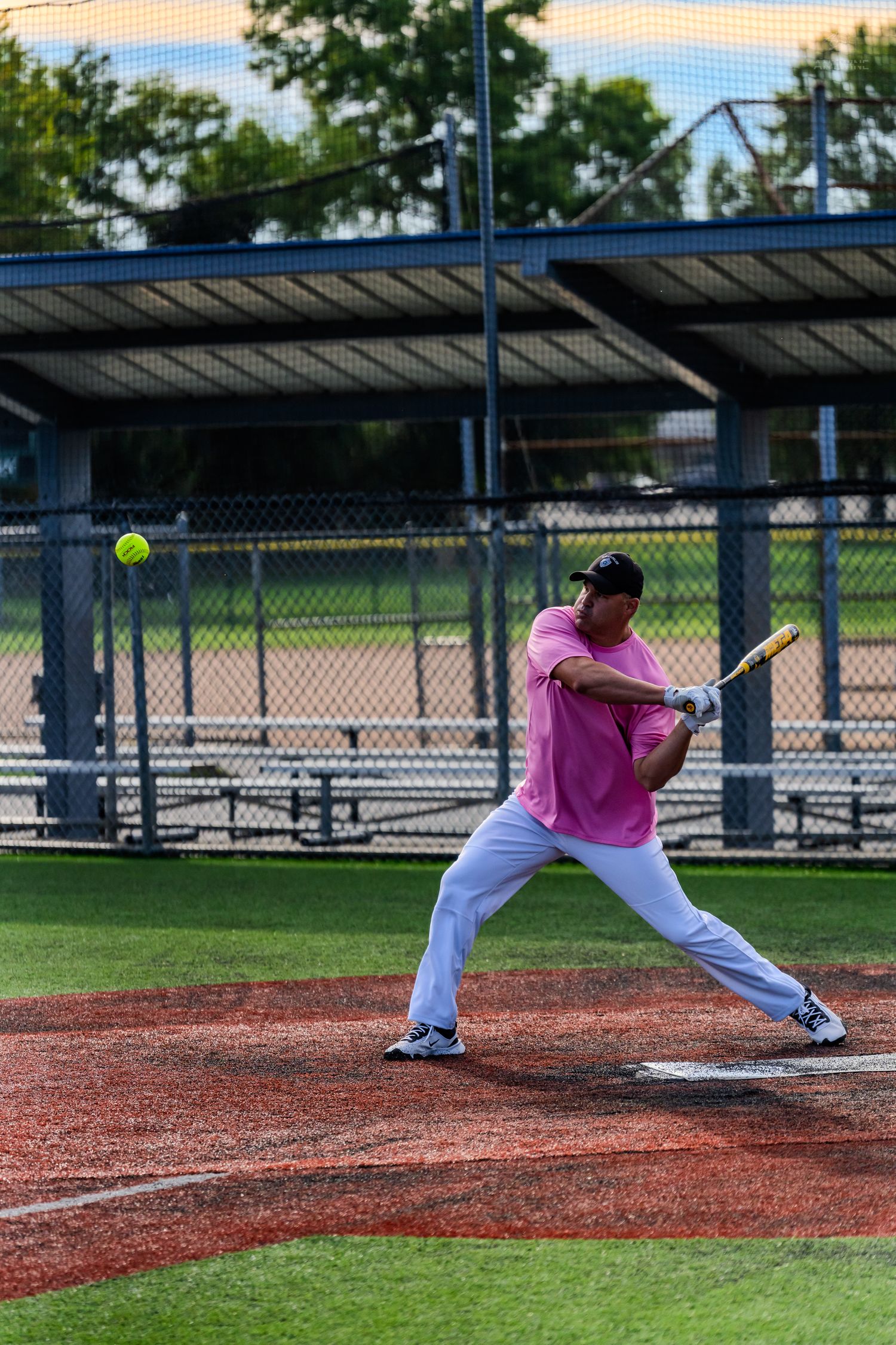A batter in a pink shirt and white pants swings at a yellow softball during a recreational league game outdoors.