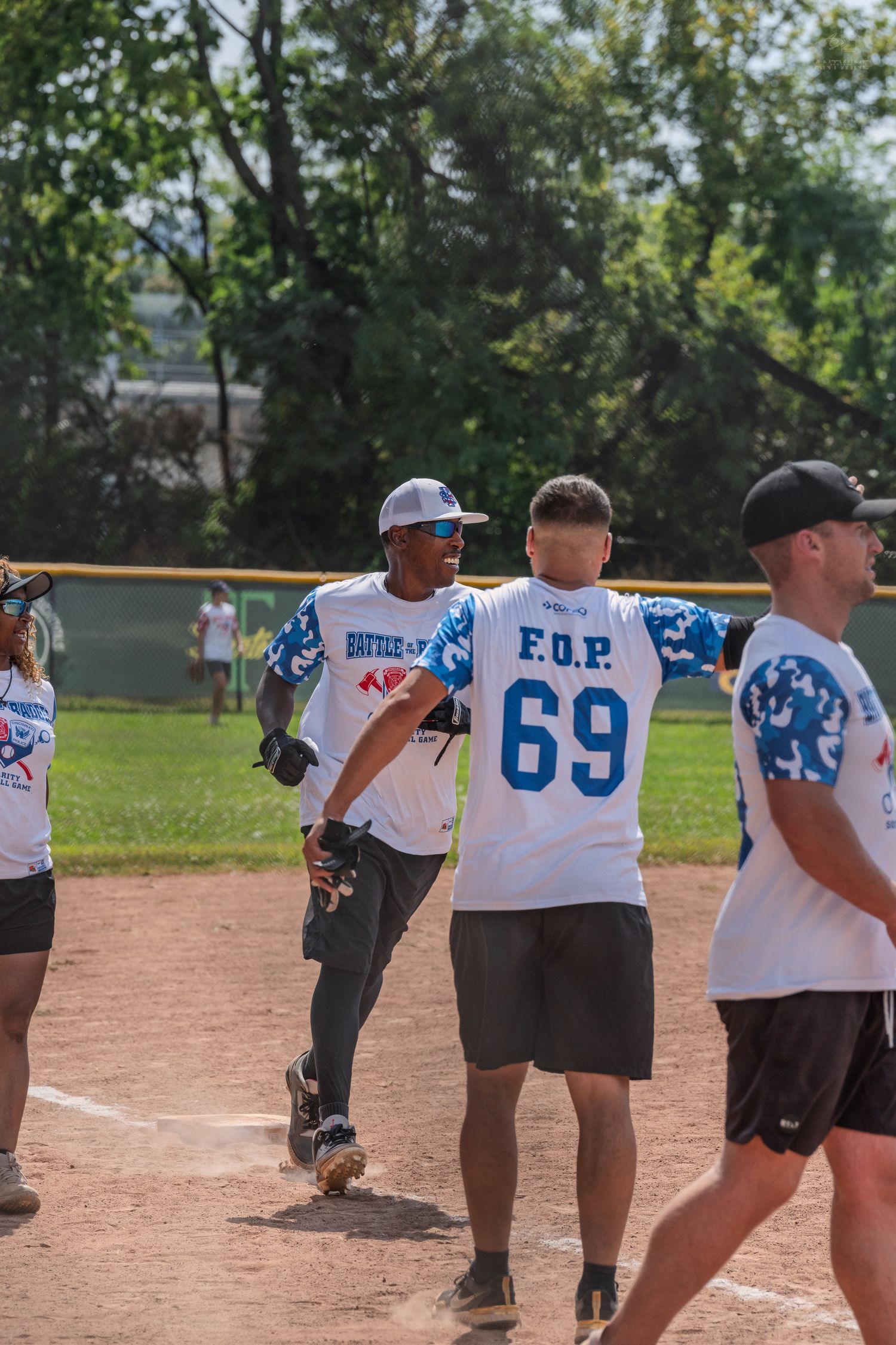Players in F.O.P. jerseys celebrate on a dirt softball field during a sunny outdoor charity game.