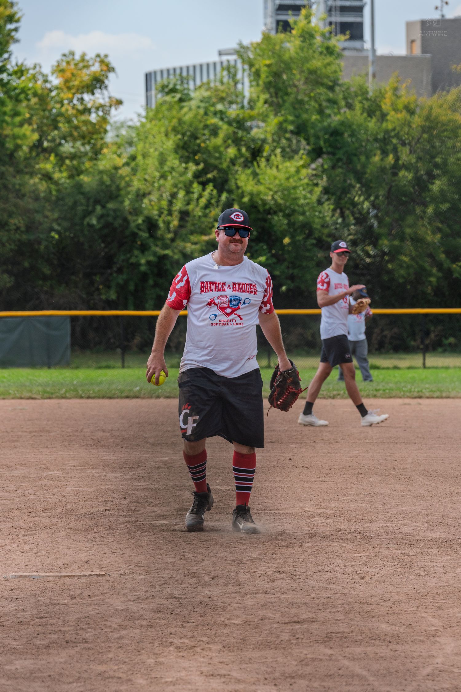 Smiling softball player in charity game jersey holds glove on dirt infield during game.