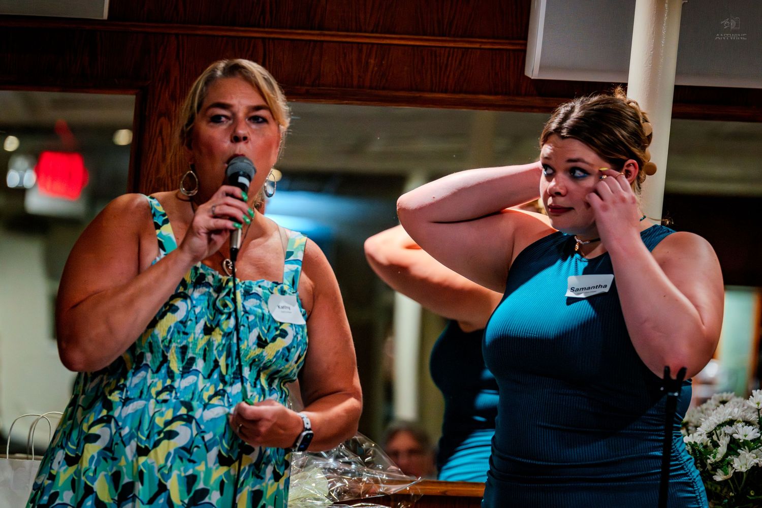 Two women at a podium, one speaking into a microphone while the other wipes tears from her eyes.