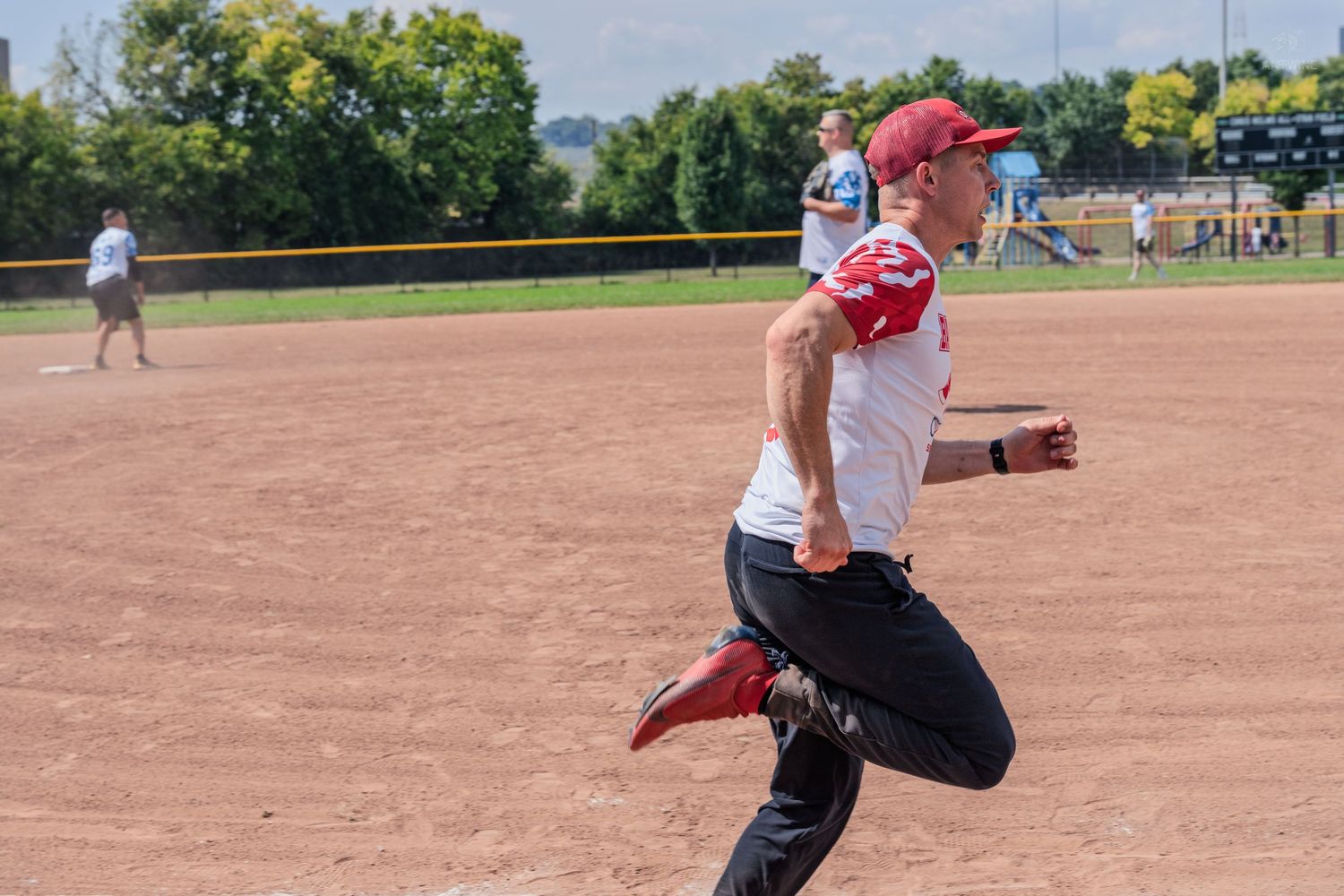 Softball player in red uniform sprinting across dirt infield during outdoor charity game.