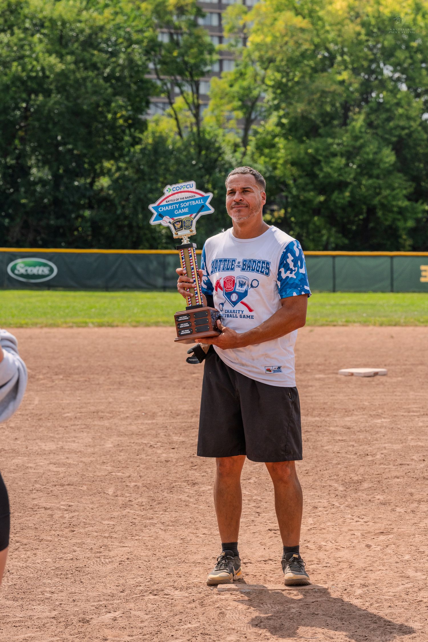 Man proudly holding a trophy on a baseball diamond during an outdoor softball tournament on a sunny day.
