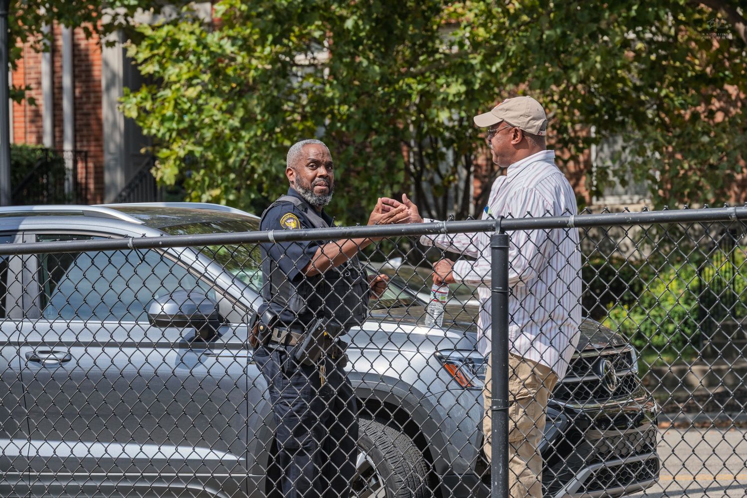 Police officer and civilian having a conversation near a chain-link fence in an outdoor parking area.