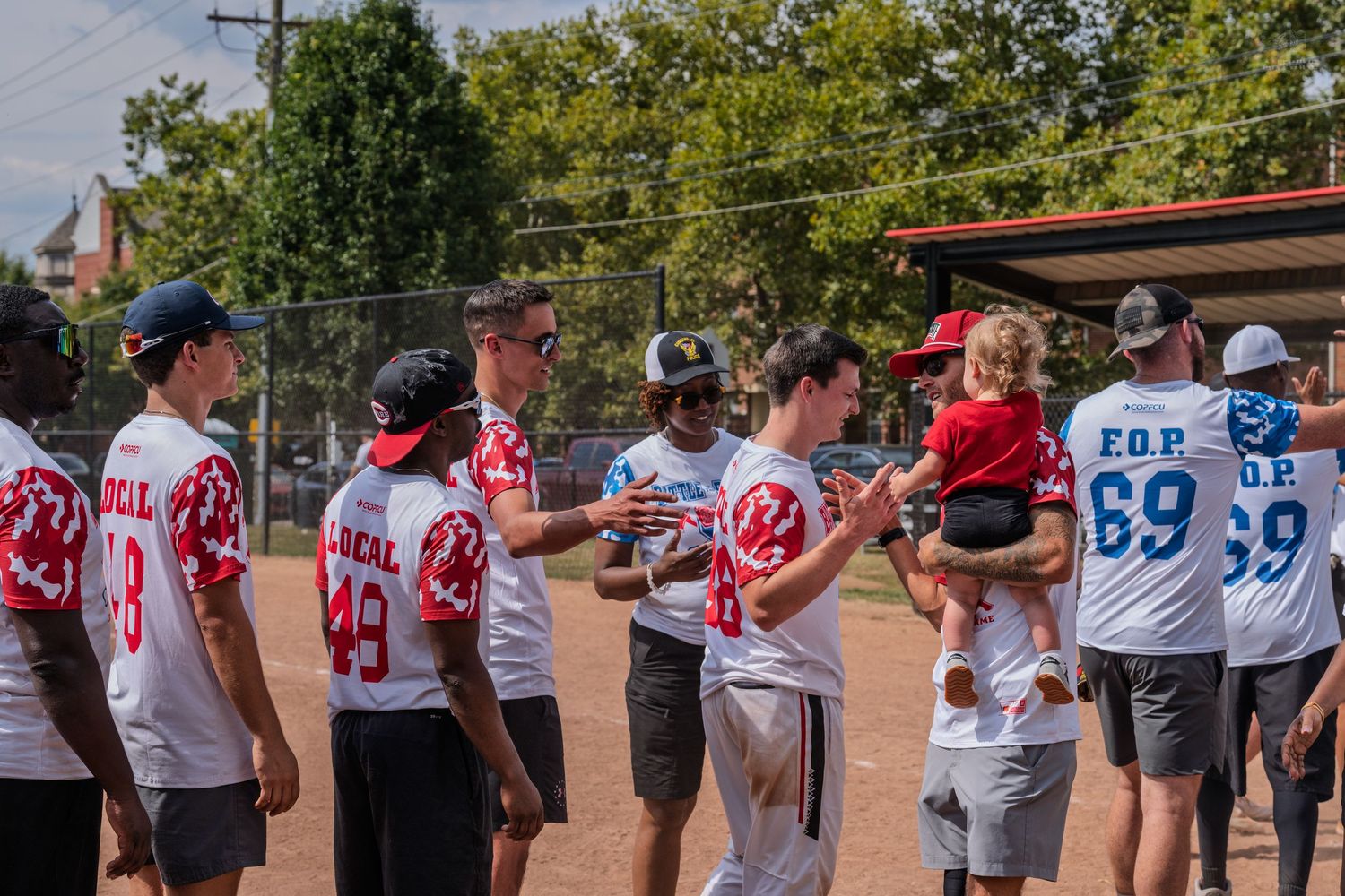 Teams in red and blue FOP softball uniforms exchanging handshakes on a baseball field after a charity game.