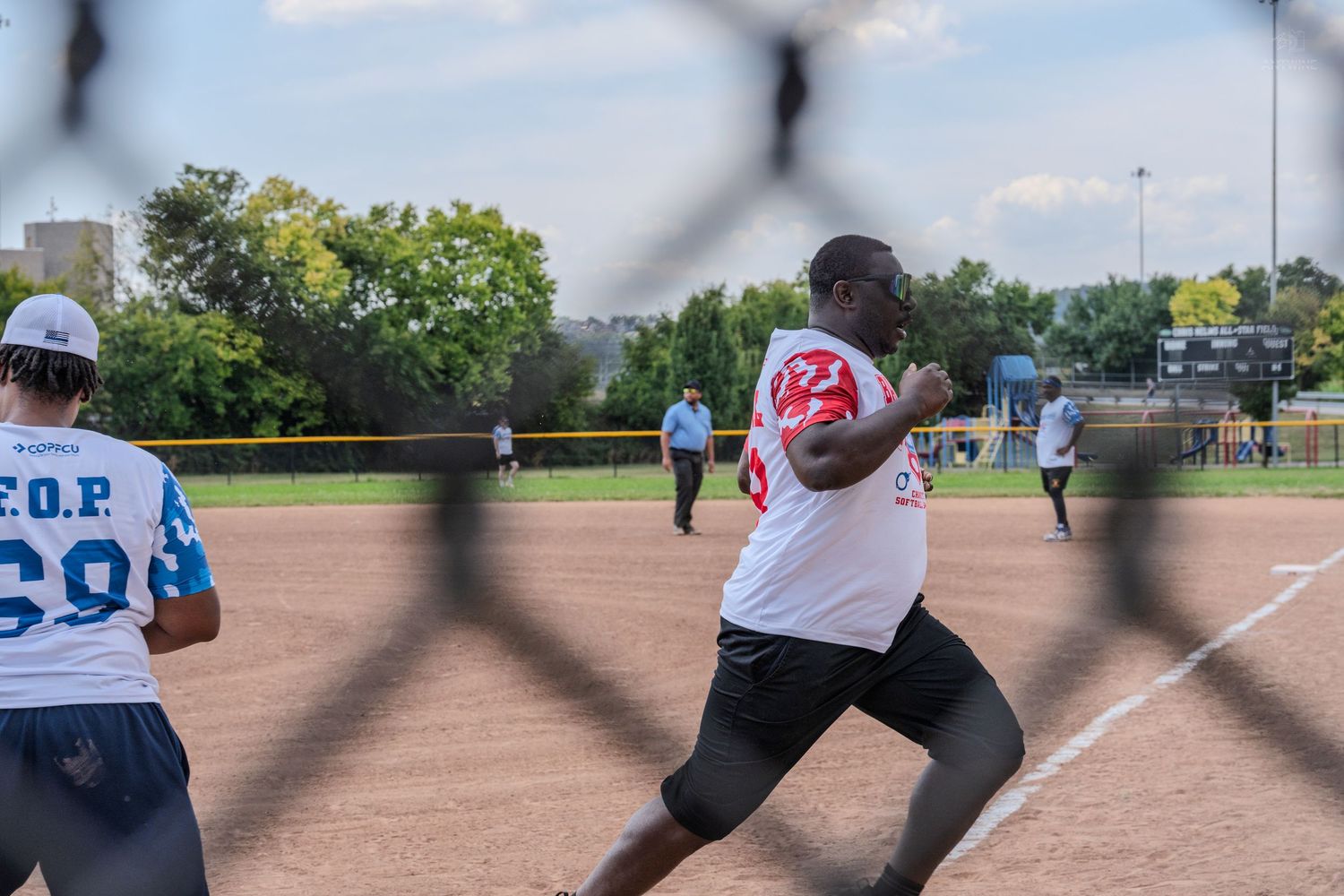 Softball player rounding a base during an outdoor game viewed through a chain-link fence.