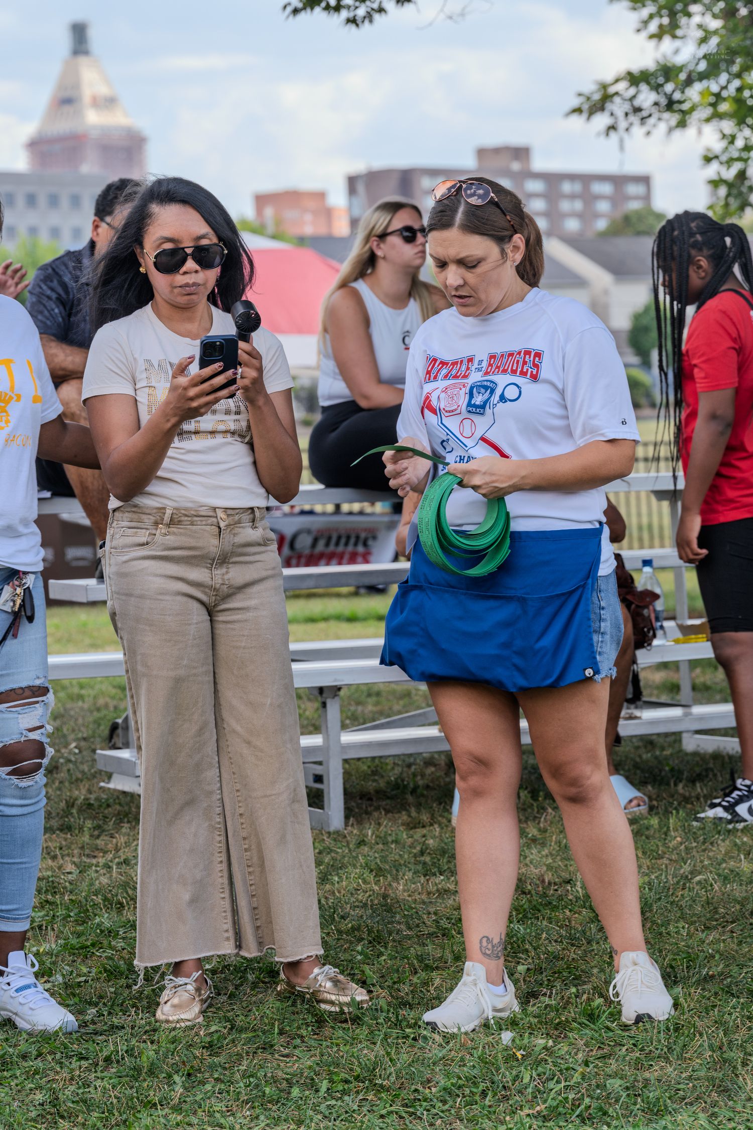 Two women standing outdoors at event, one checking phone, one holding hat and frisbee.