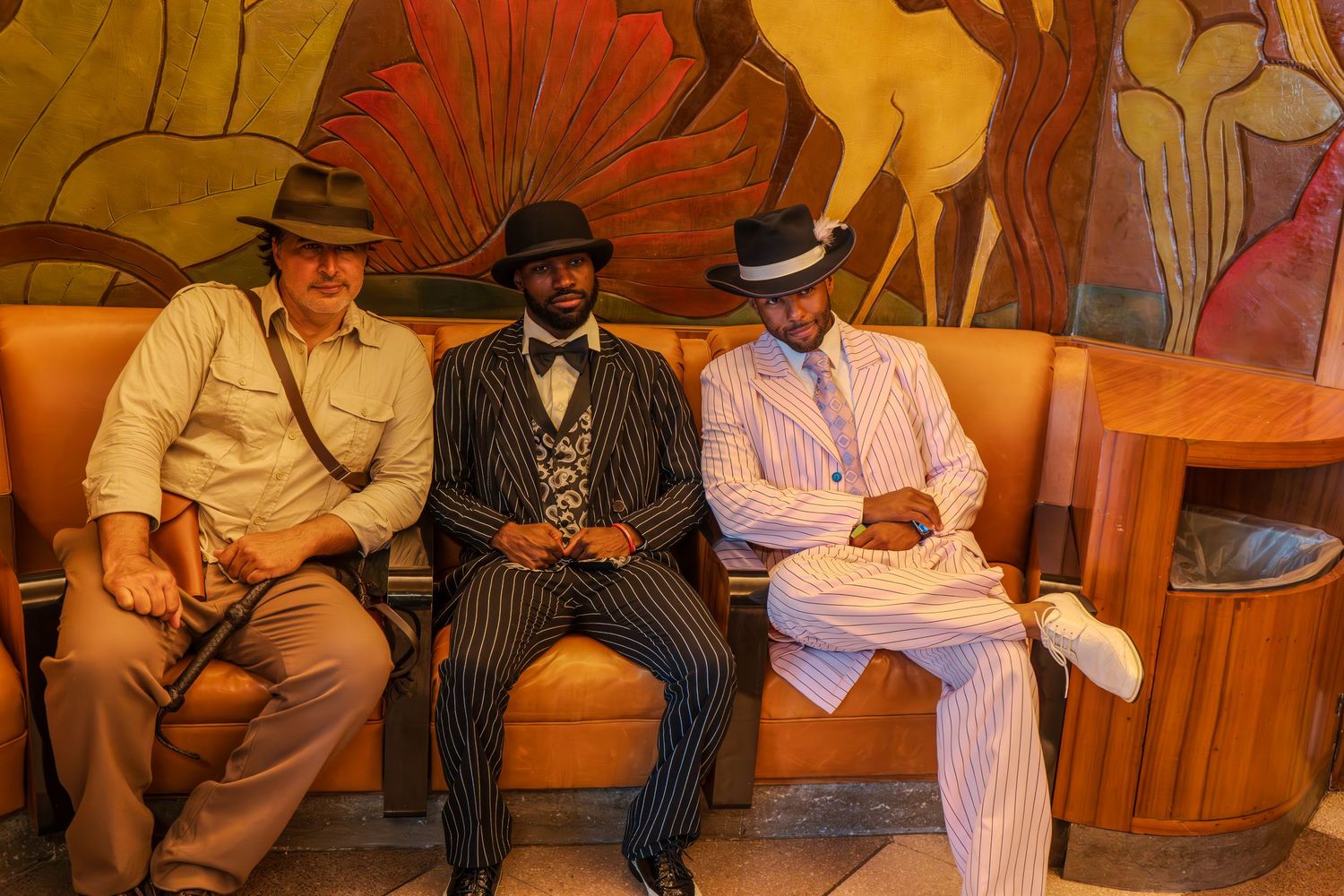 Three men in vintage 1920s-style suits and fedora hats sitting on a bench before a colorful mural.