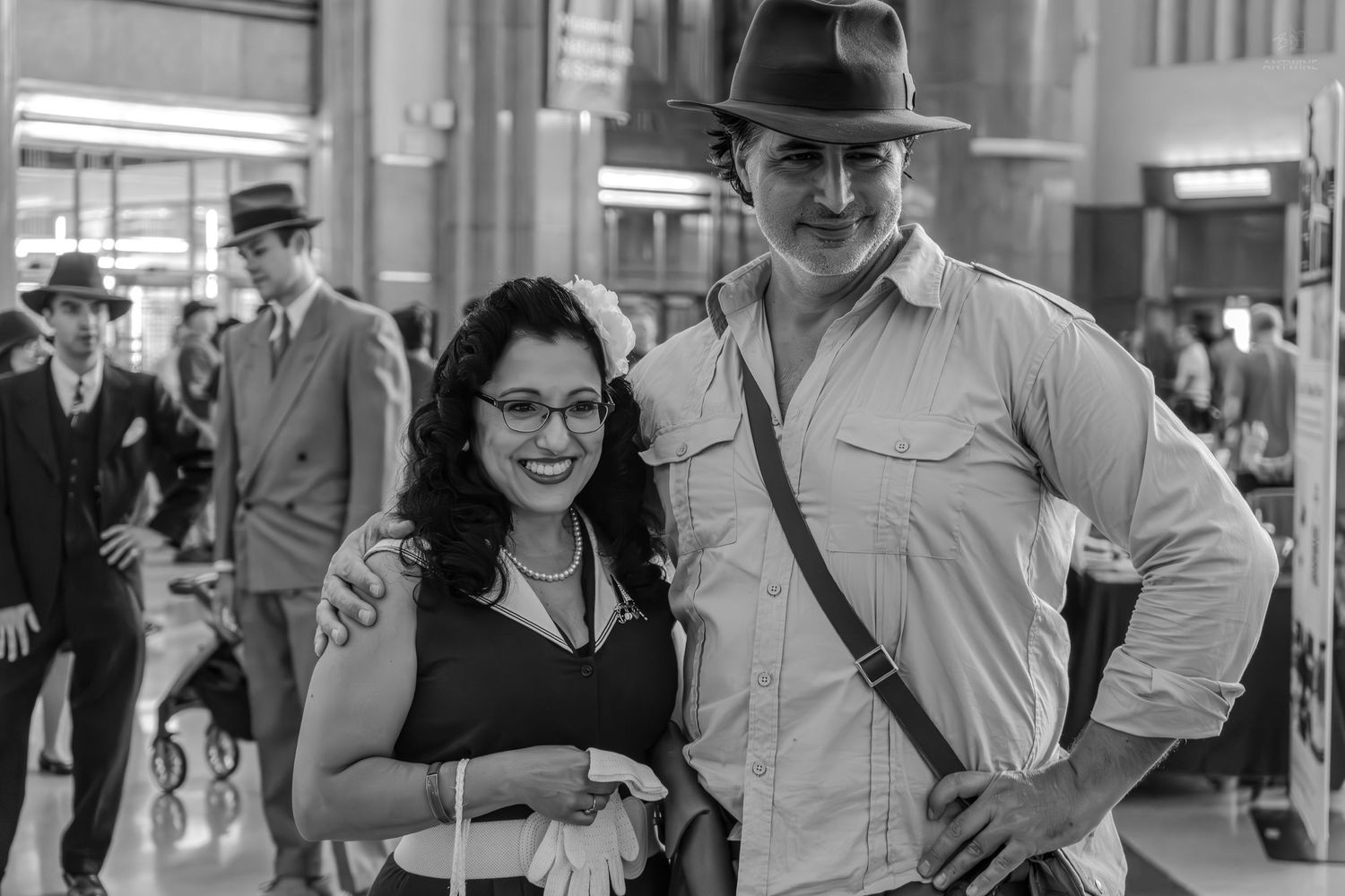 Black and white photo of a smiling couple in vintage 1940s costumes at a retro-themed event.