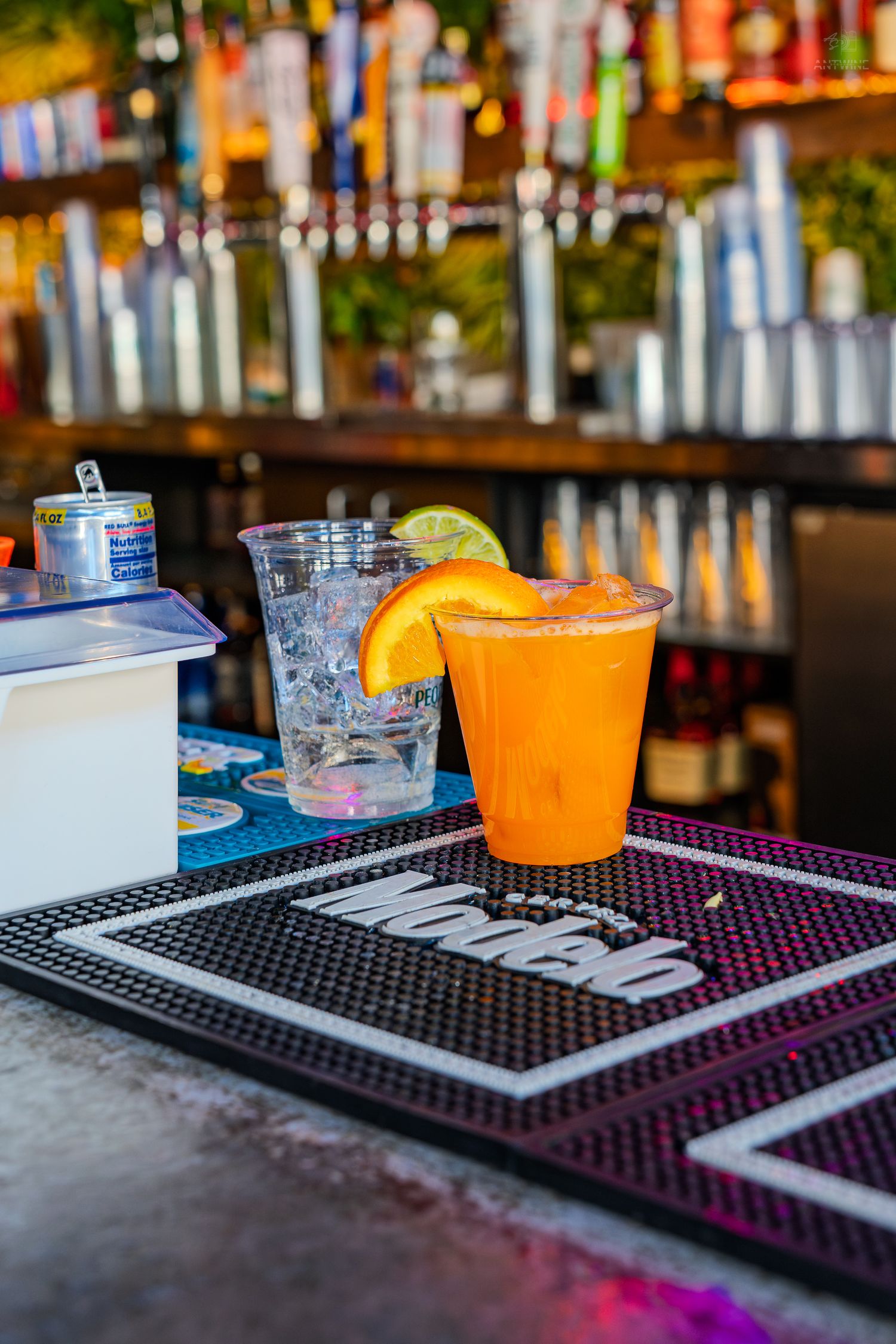 Two cocktails on a Modelo bar mat, featuring an orange drink garnished with citrus and a clear drink with ice.