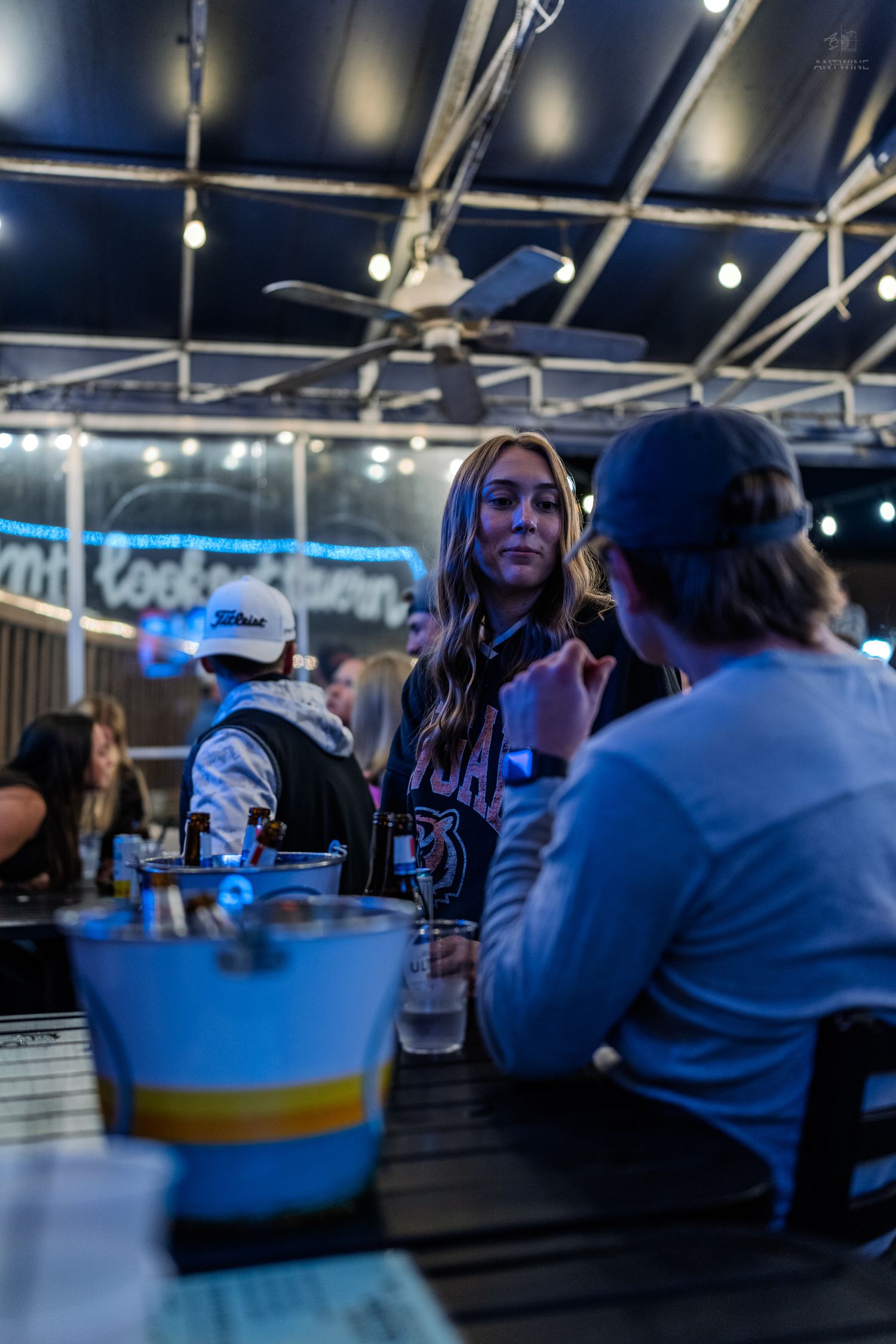 Woman in graphic tee chatting with friend at a lively outdoor bar patio with string lights at night.