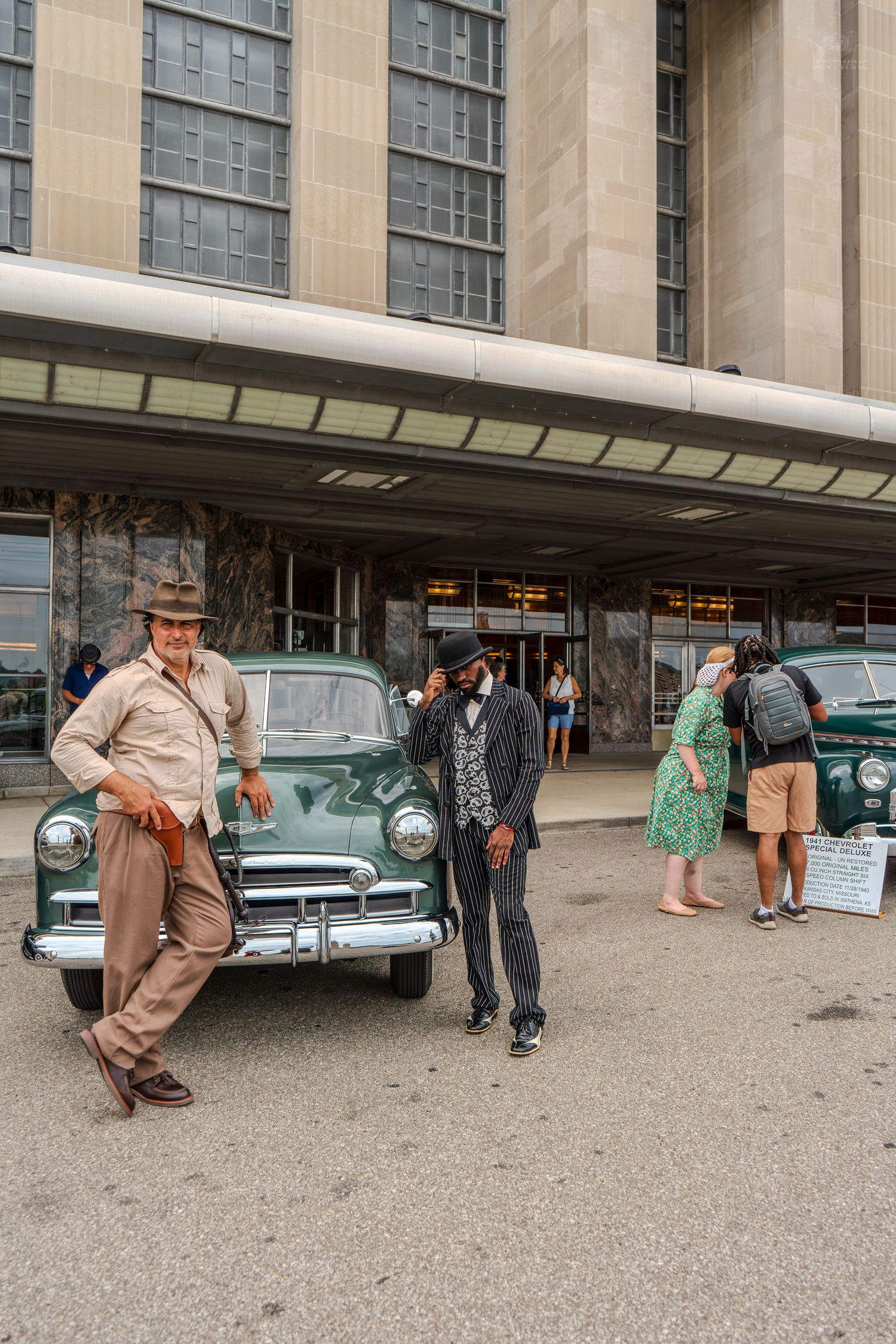 Two men in vintage attire pose beside a classic green car outside a large Art Deco building.