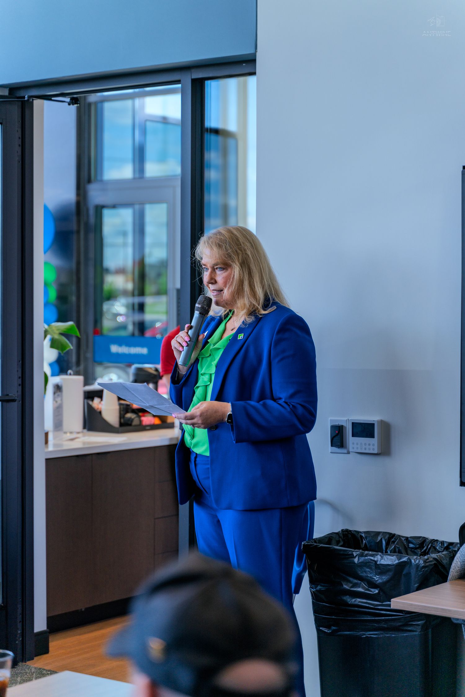 Woman in blue blazer and green shirt speaks at podium with microphone at indoor corporate event.