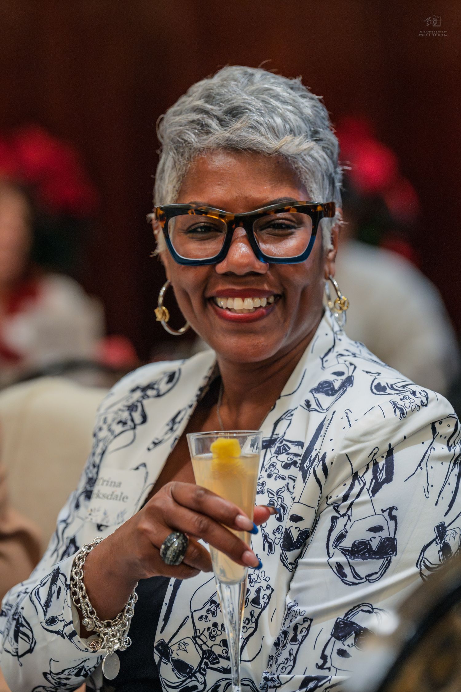Smiling woman with gray hair and glasses holding a cocktail at an event.
