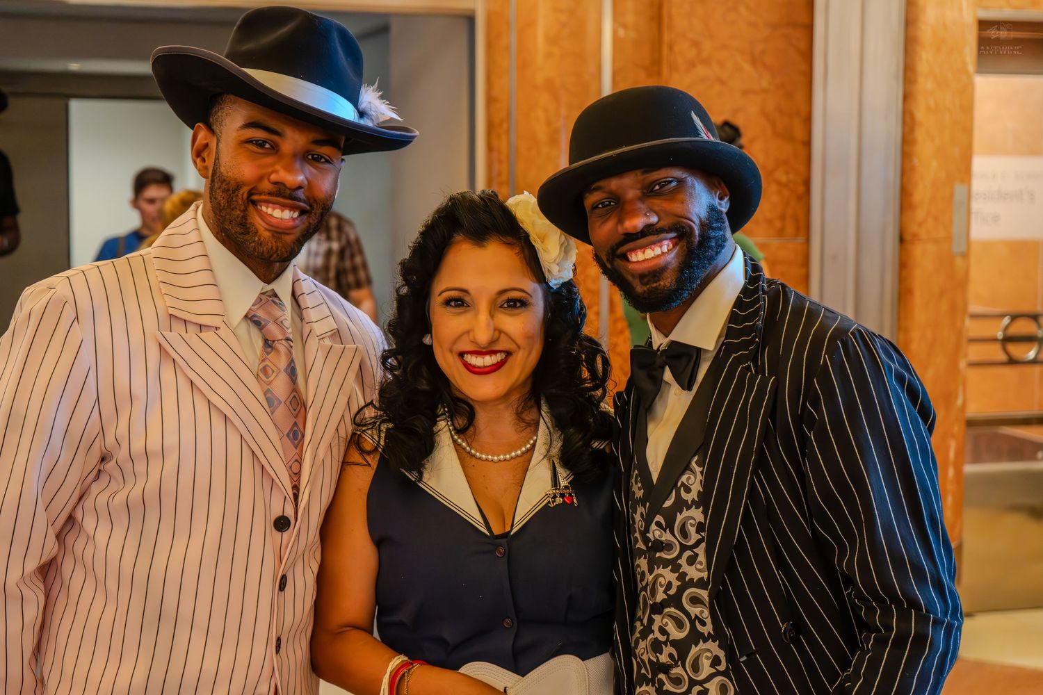 Three elegantly dressed people posing together at a formal event, wearing vintage-style suits and hats.