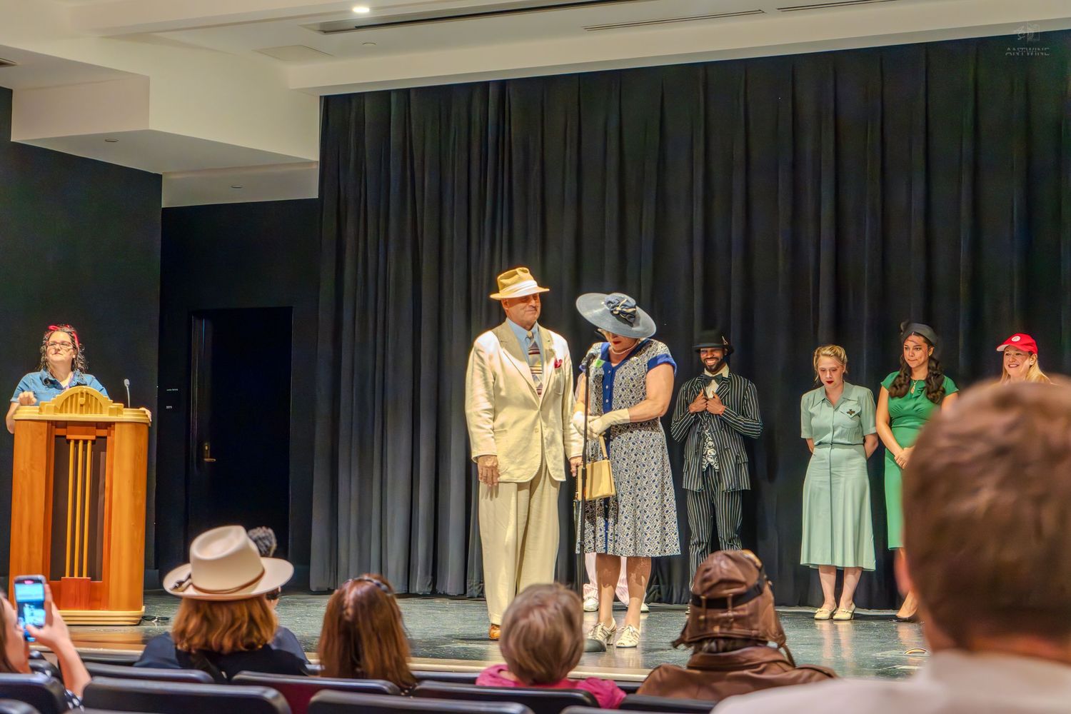 Actors in vintage 1940s costumes perform on stage during a theatrical production with an audience watching.
