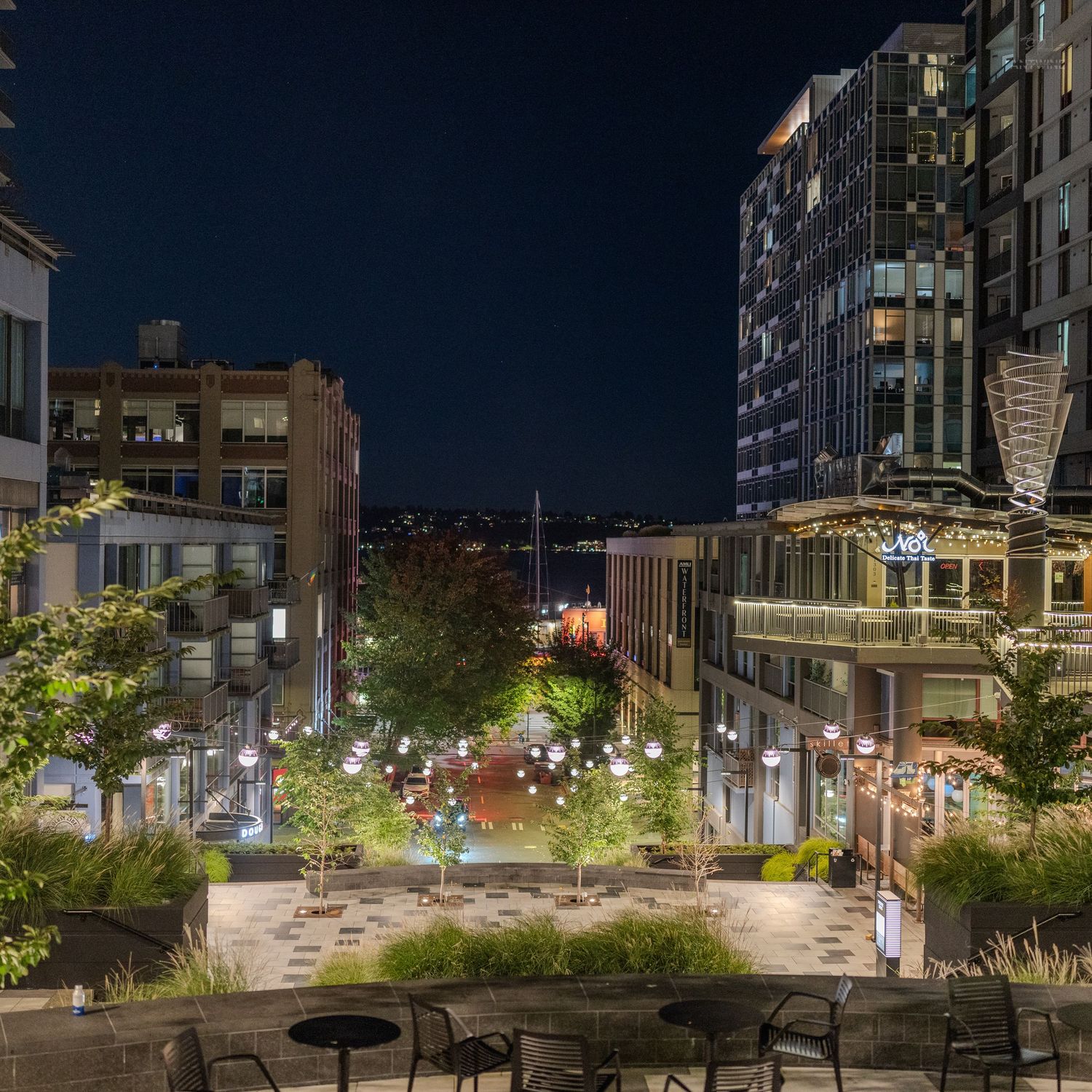 Nighttime urban plaza with string lights, trees, and modern high-rise buildings illuminated against a dark sky.