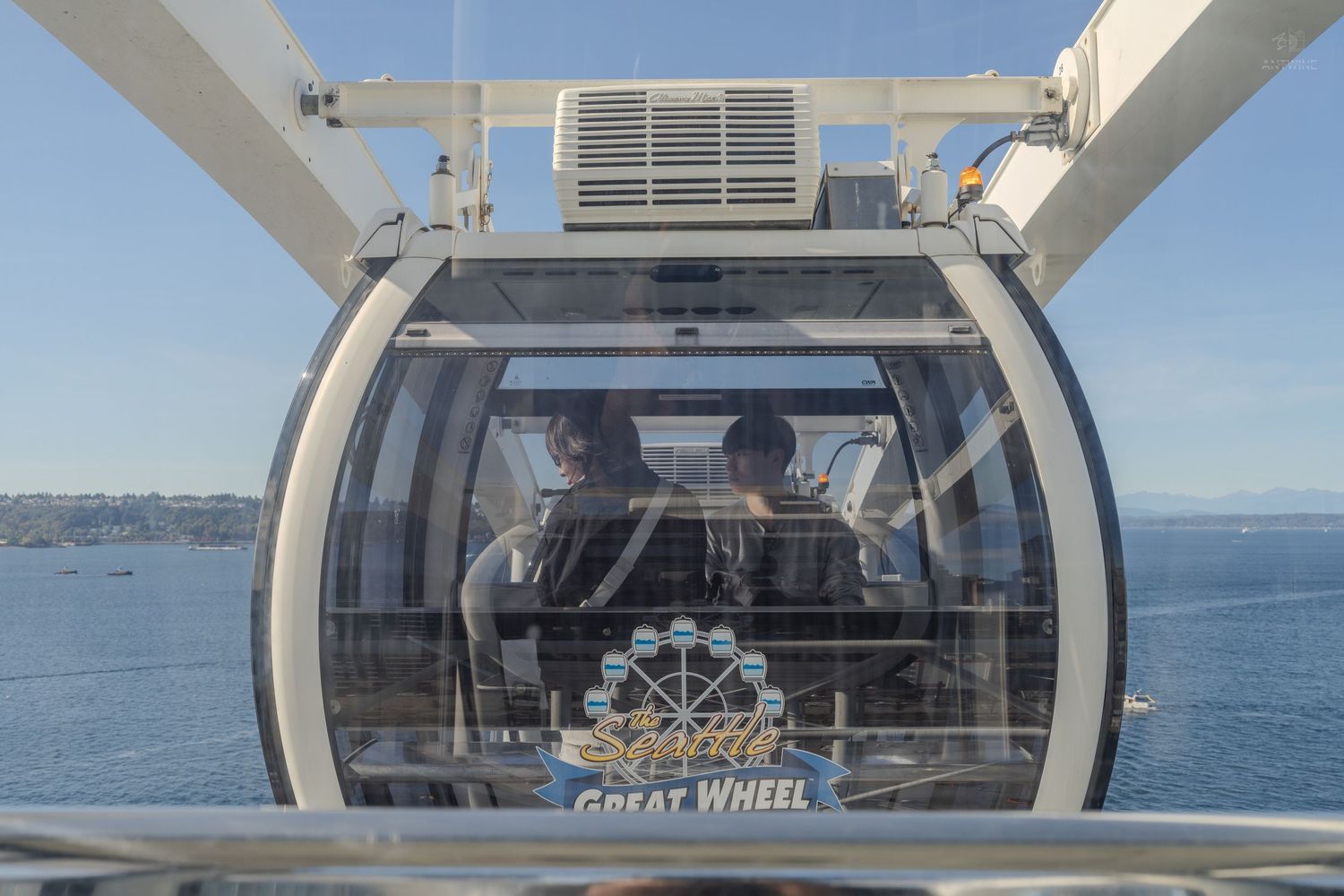 Two passengers ride inside a glass gondola cabin on the Seattle Great Wheel Ferris wheel overlooking the bay.