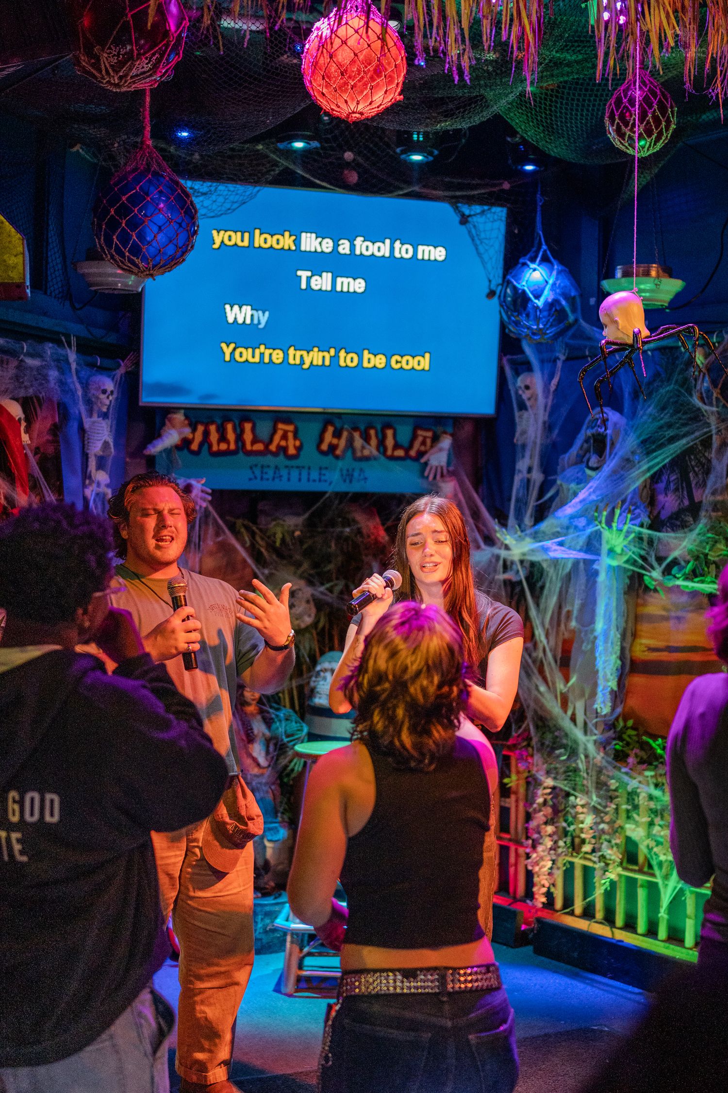 Young woman sings karaoke at Hula Hula bar with Halloween decorations and lyrics displayed on screen behind her.