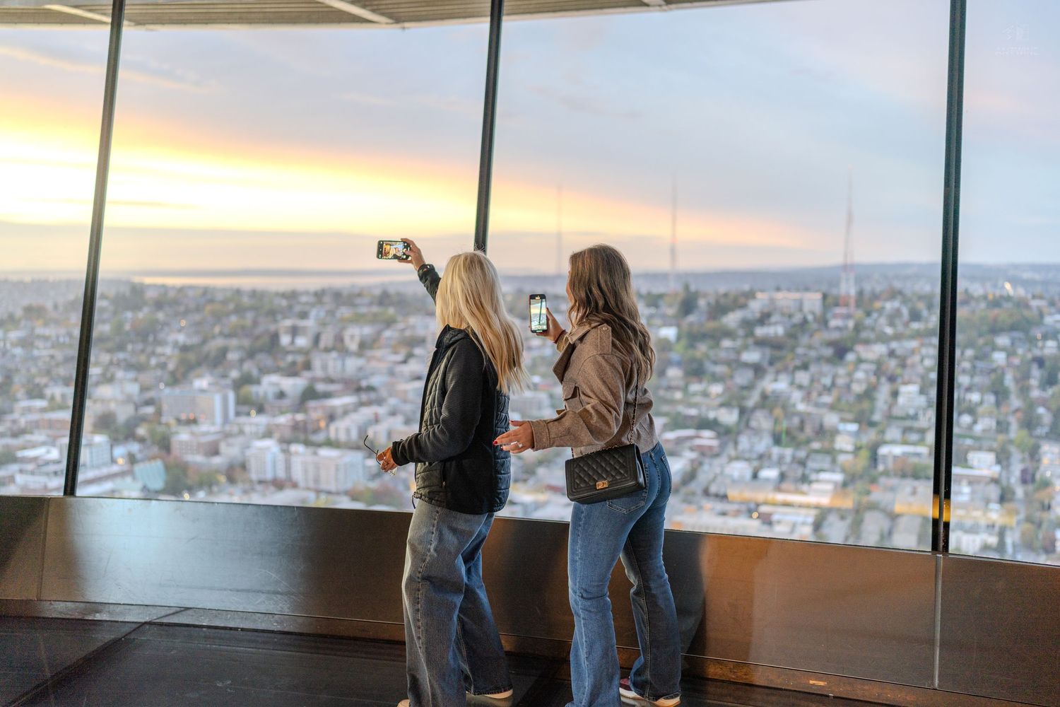 Two women take selfies at a high-rise observation deck overlooking a city at sunset.