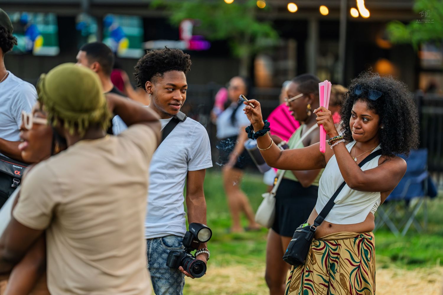 Group of young adults socializing at outdoor festival, woman holds pink drink while man with camera smiles nearby.