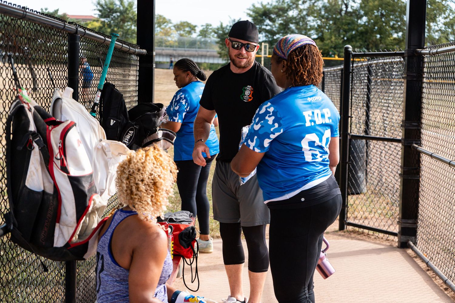 Group of people in blue jerseys socializing near a chain-link fence at an outdoor sports facility.