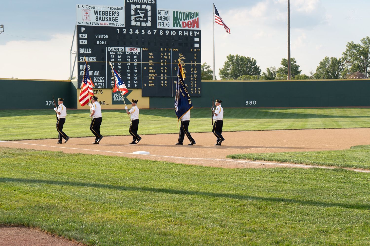 Color guard performing at a minor league baseball stadium during a pre-game ceremony with a scoreboard in the background.