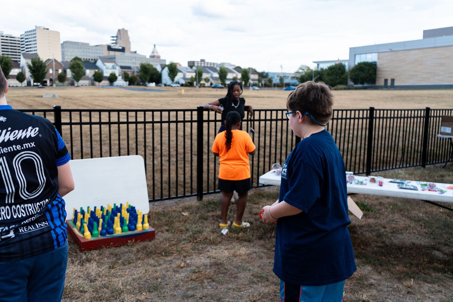 People gathered outdoors near fence with colorful artwork display on field.