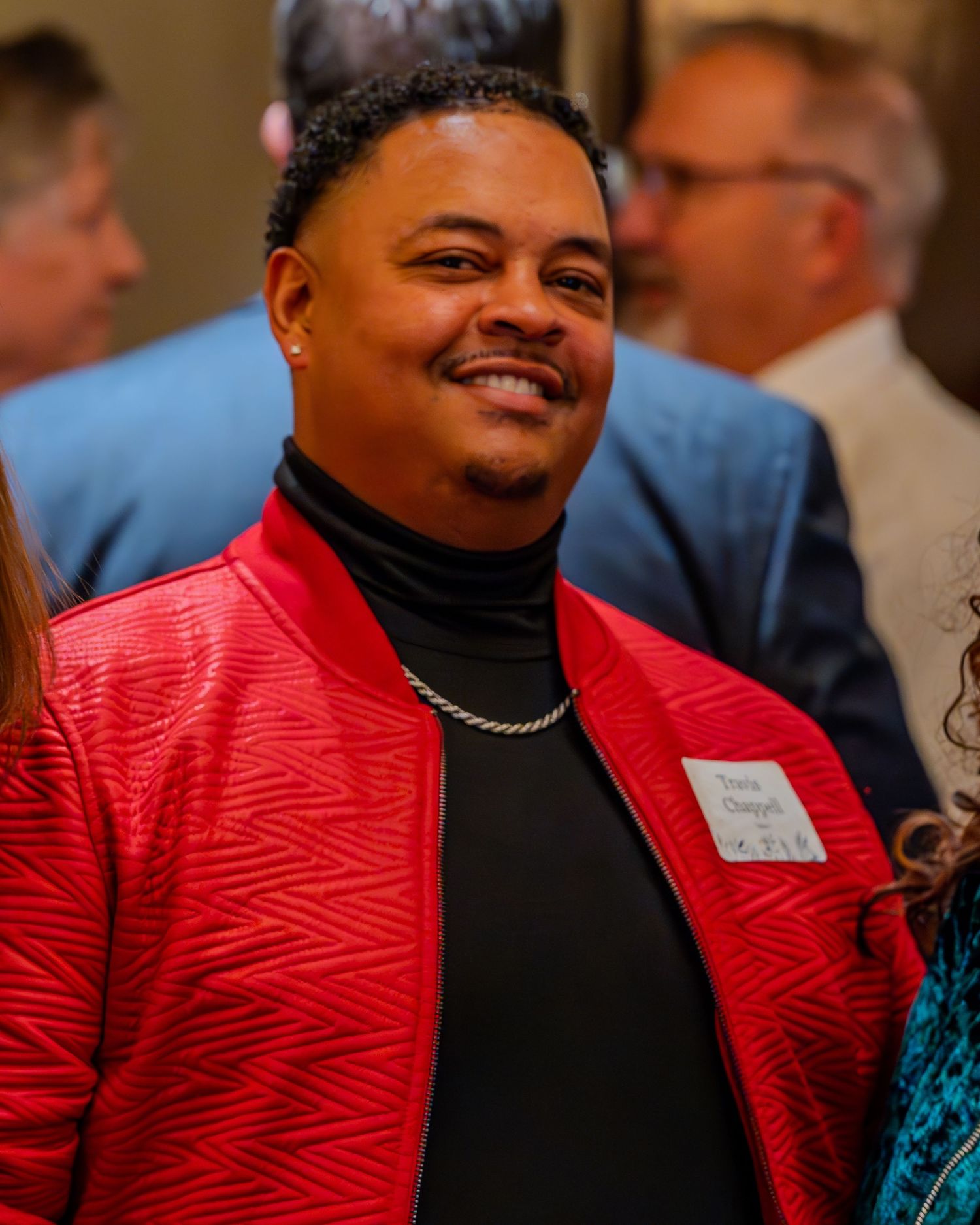 Smiling man in red jacket and black turtleneck with silver chain at a social event.
