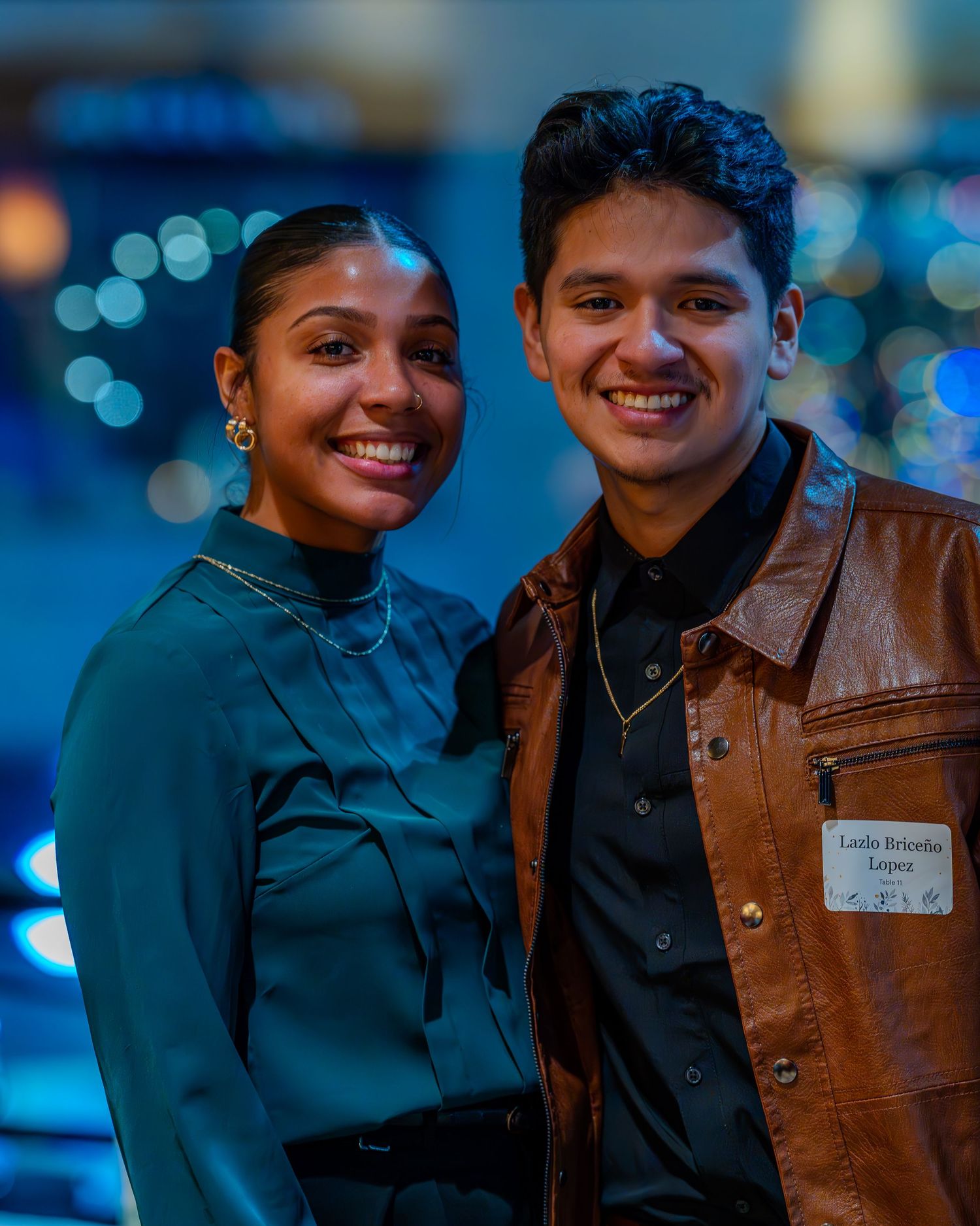 Two smiling young adults pose together at a formal event, man wearing brown leather jacket with name tag.