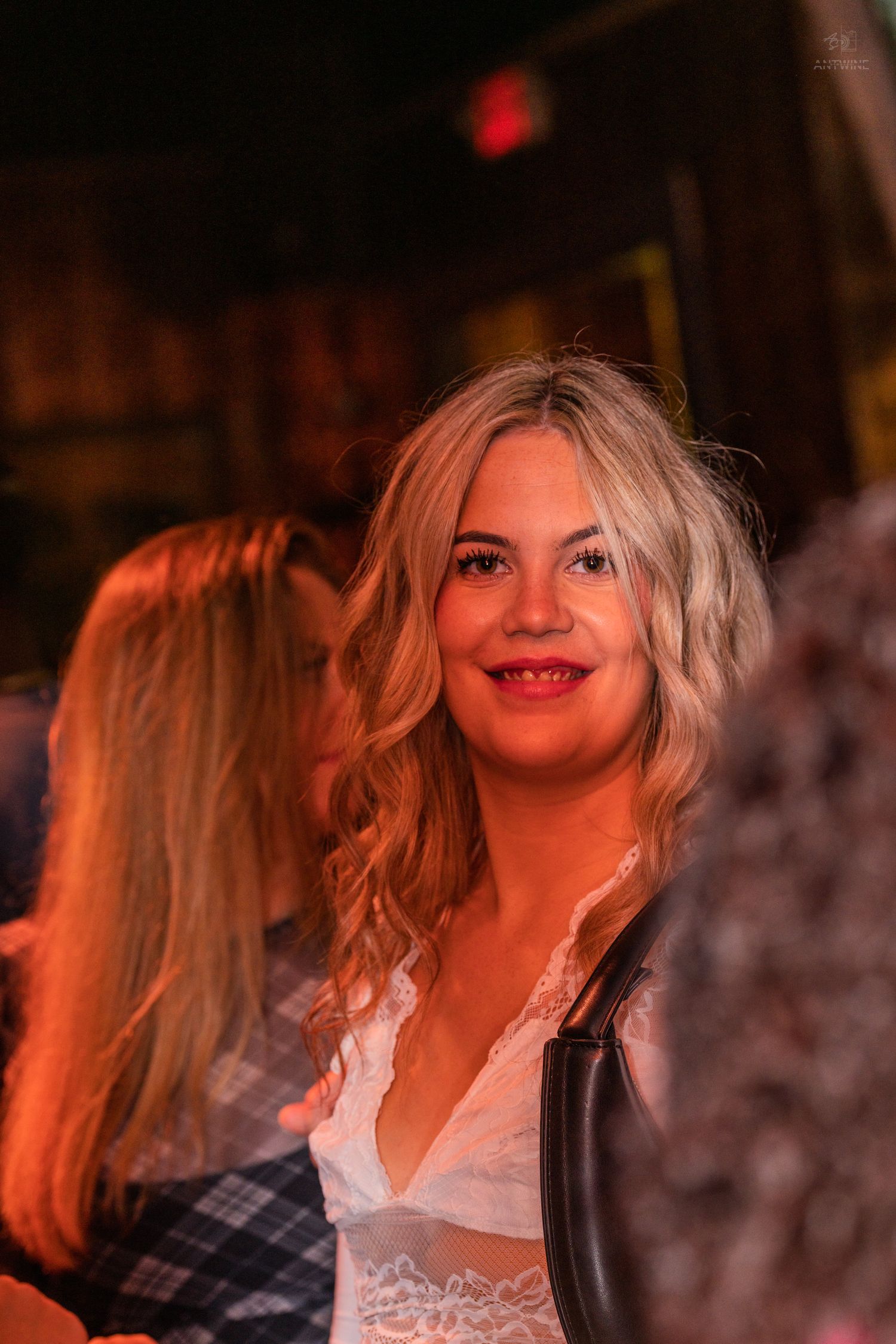 Smiling blonde woman in white lace top at a dimly lit nightclub or bar event.