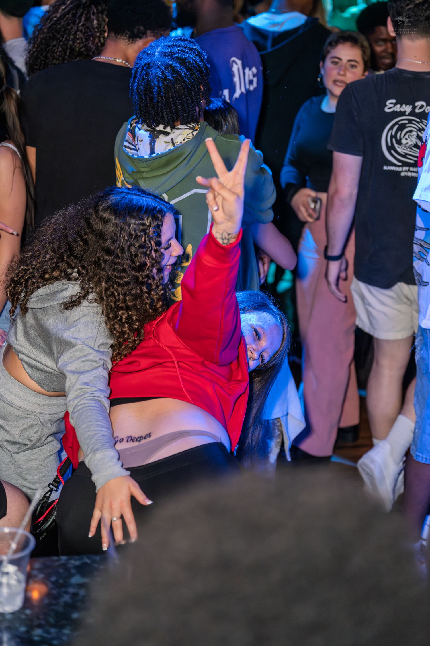 Young woman in red hoodie dancing and flashing peace sign in a crowded nightclub.