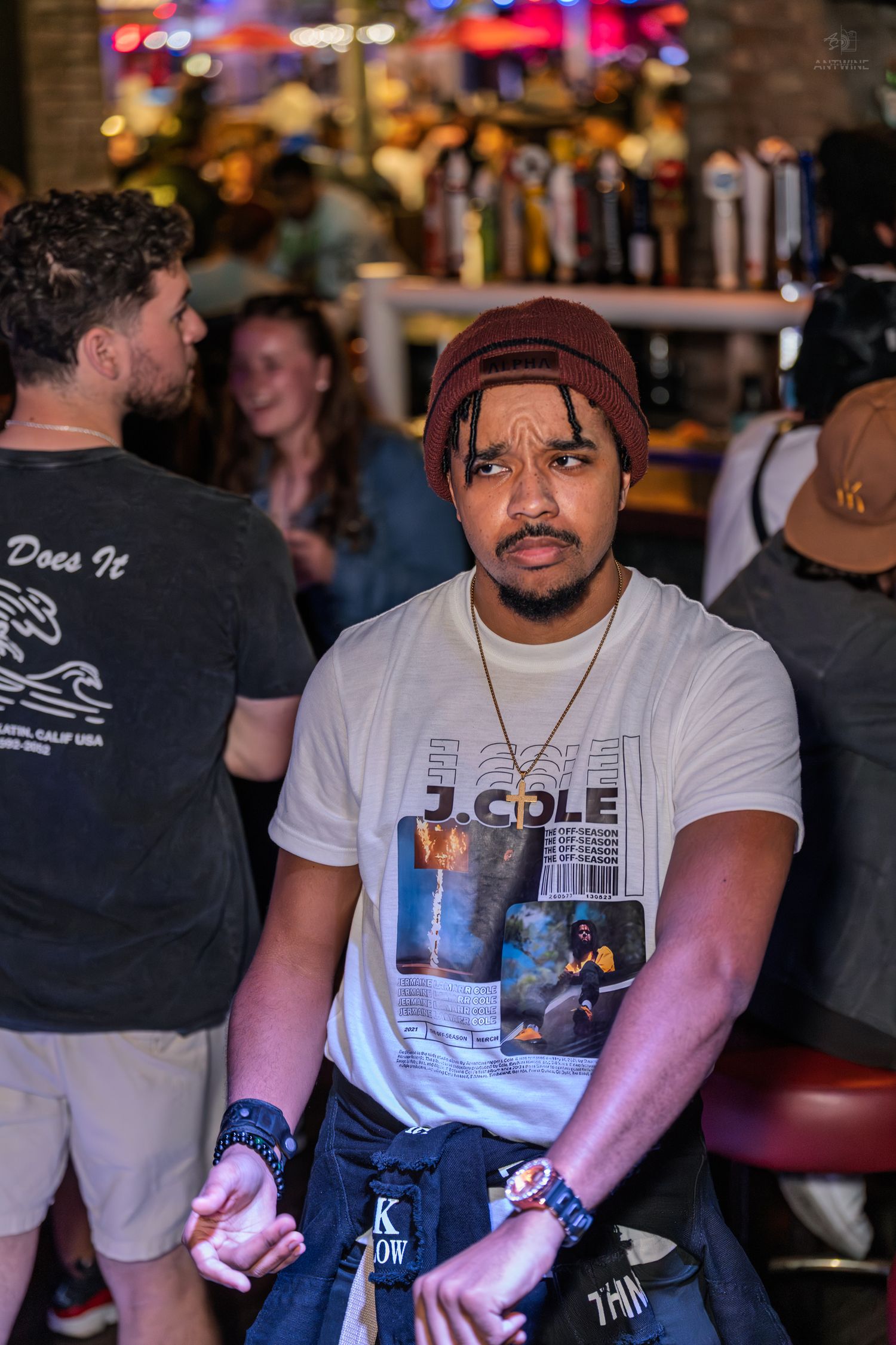 Young man in J. Cole graphic tee and burgundy beanie at a crowded bar.