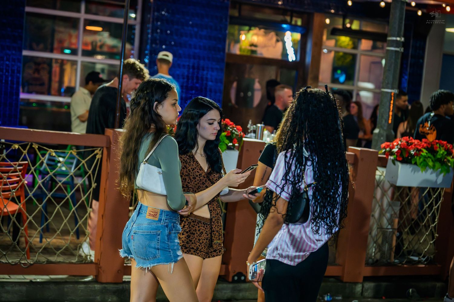 Three young women in casual outfits chat outside a restaurant with string lights at night.