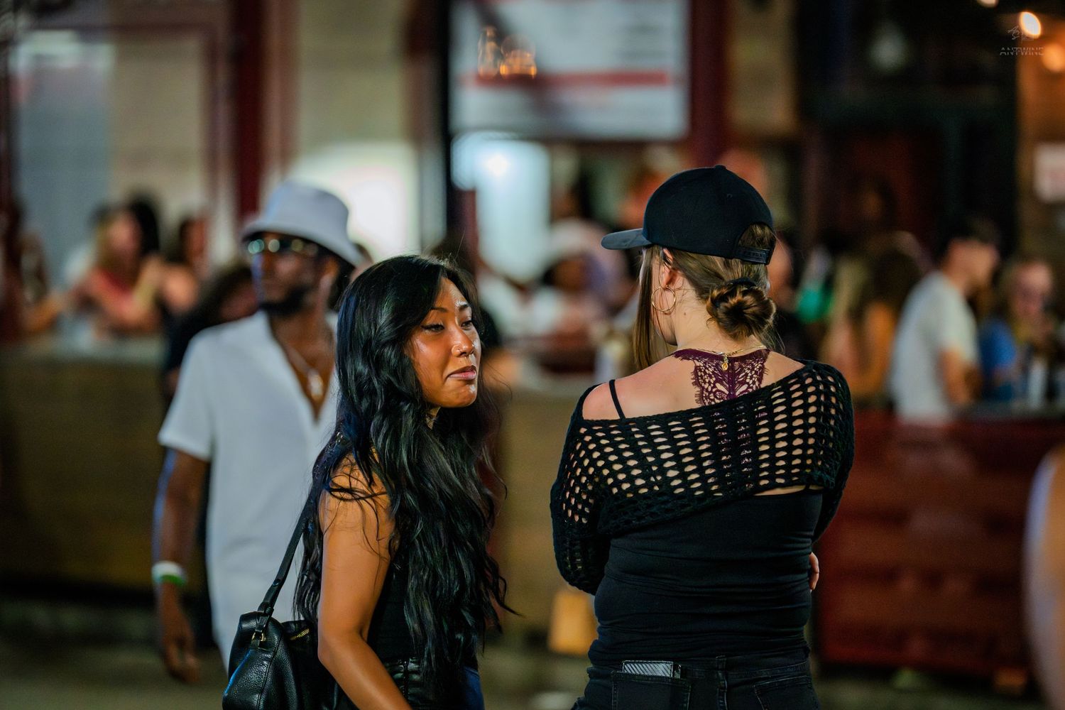 Two women in black outfits converse outside a busy nightlife venue at night.