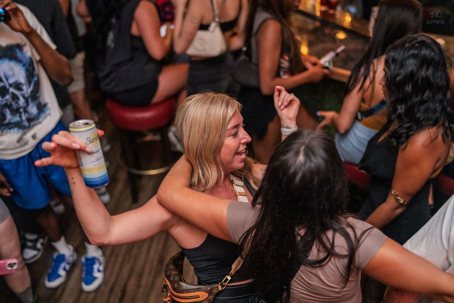 Blonde woman raising a canned drink while dancing and laughing with friends at a bar.